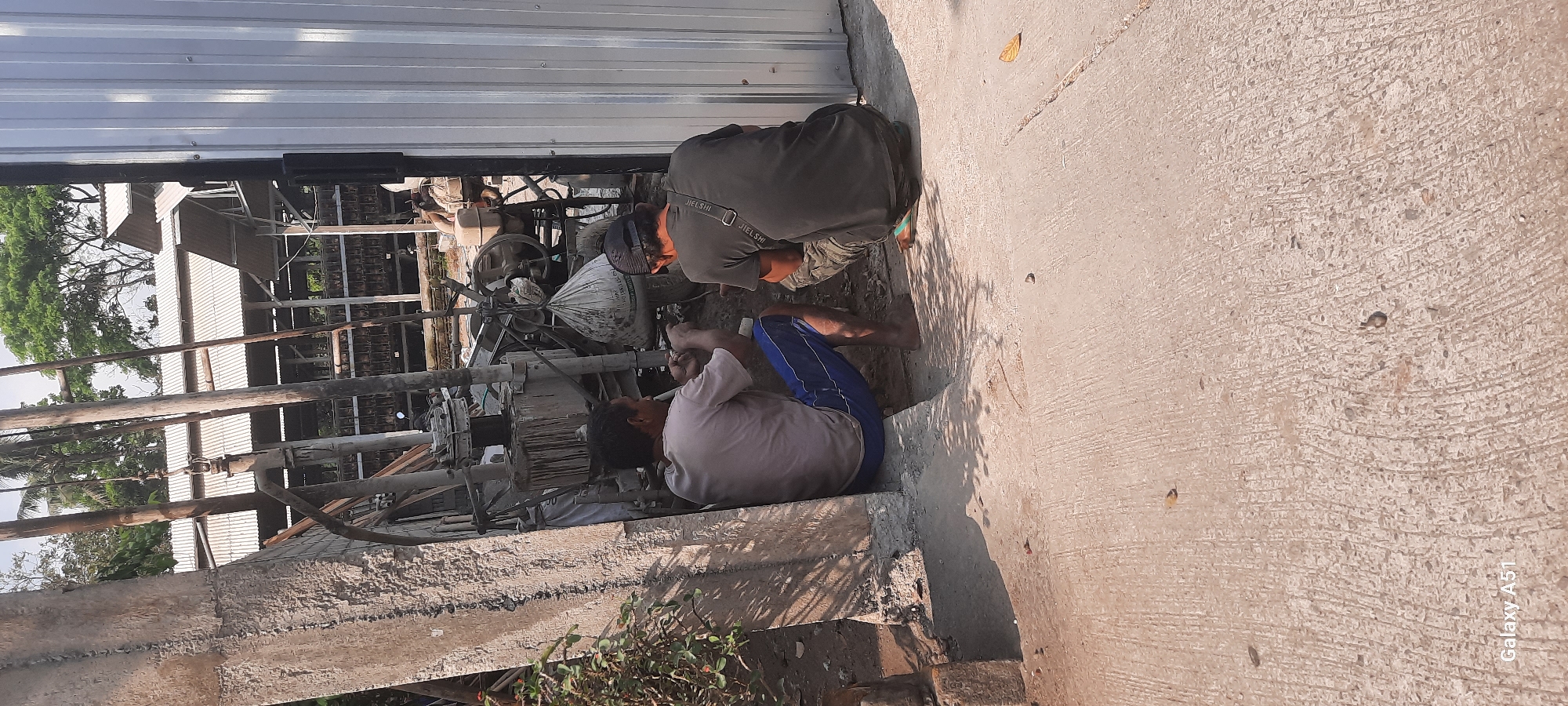 Two men are working on equipment in an outdoor industrial area in Ploso, Indonesia. The man on the left, wearing a light-colored t-shirt and blue shorts, is sitting and appears to be manipulating a tool. The man on the right, wearing a dark t-shirt and camouflage shorts, is crouching and also engaged with the equipment. They are positioned near a large concrete structure on the left and a corrugated metal wall on the right. In the background, there is a structure with a metal roof, trees, and what appears to be machinery, including a large engine and a suspended white bag. The ground is paved with concrete.