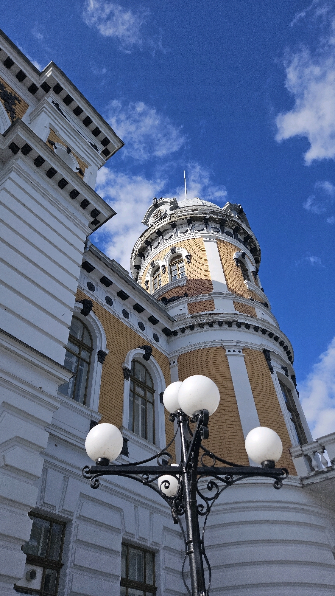 A low-angle shot captures a section of a yellow brick building with white architectural details, including a round tower topped with a dome, under a bright blue sky with scattered white clouds. In the foreground, a black ornate lamppost with four white spherical lights is positioned slightly to the right. The lamppost's design includes decorative scrollwork. The building features arched windows and elaborate cornices. This architecture is characteristic of the Ulyanovsk Regional Museum of Local Lore, named after Ivan Goncharov, located in Ulyanovsk, Russia. The scene is static, with no people or animals visible. The lighting suggests it is daytime.