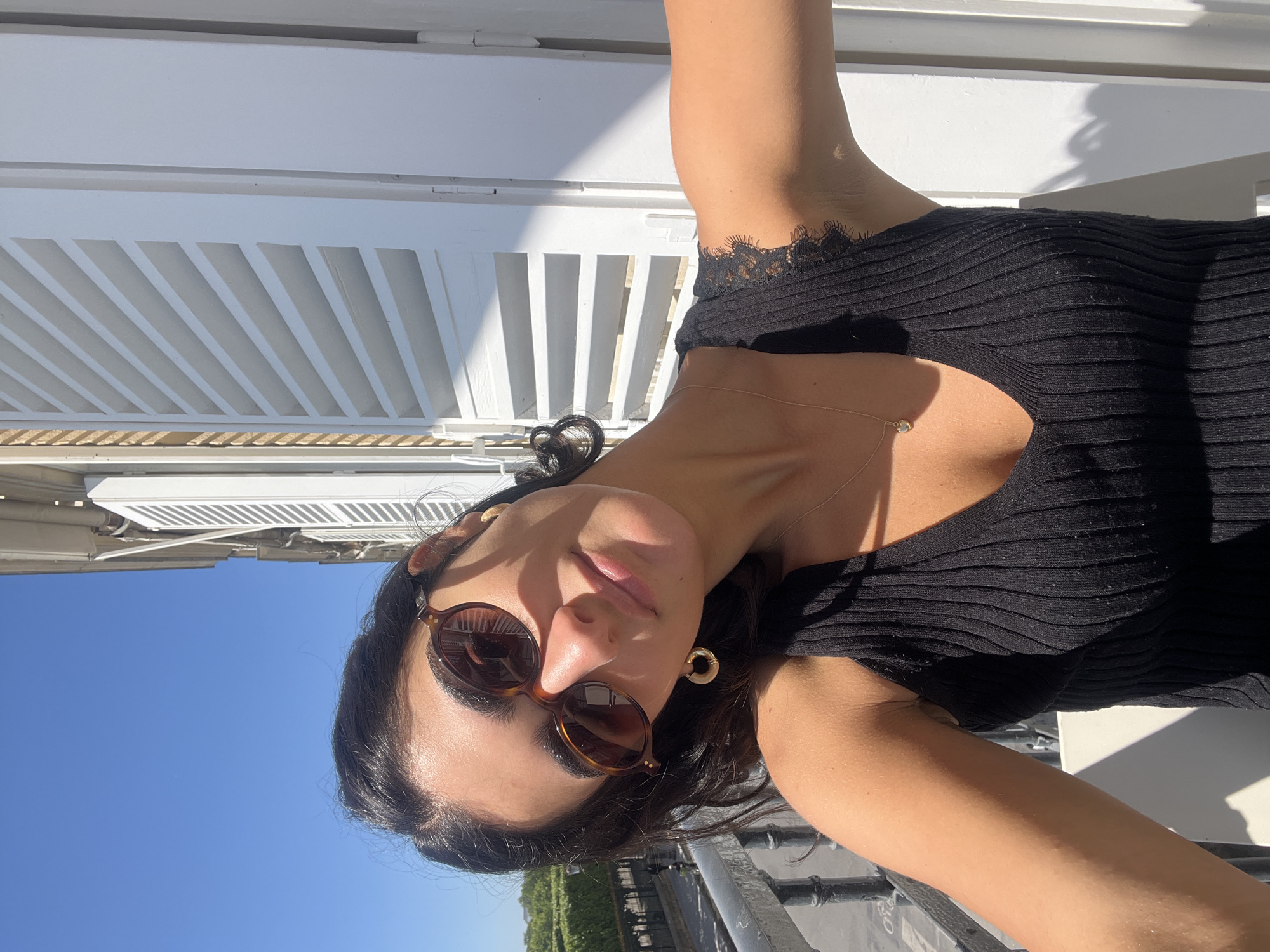 A young woman is captured in a selfie taken outdoors, likely on a balcony or terrace in Paris, France. The image is rotated 90 degrees counter-clockwise. She looks directly at the camera with a relaxed expression and a slight smile, her head tilted. She wears stylish tortoise-shell sunglasses, which cast shadows on her face, and a black ribbed top with delicate lace trim along the neckline or shoulder. A thin, delicate necklace with a small pendant and a circular gold earring are also visible.
The setting is brightly lit by direct sunlight, creating strong shadows on both the subject and the background. Behind her, prominent white louvered shutters dominate the upper part of the background, showing alternating patterns of light and shadow from the sun. A clear, bright blue sky is visible, suggesting good weather. In the lower part of the frame, a metal railing is present, and in the distance, subtle greenery from trees and perhaps urban structures can be discerned, consistent with a city environment. The overall impression is a sunny daytime scene. No discernible text is visible in the image.