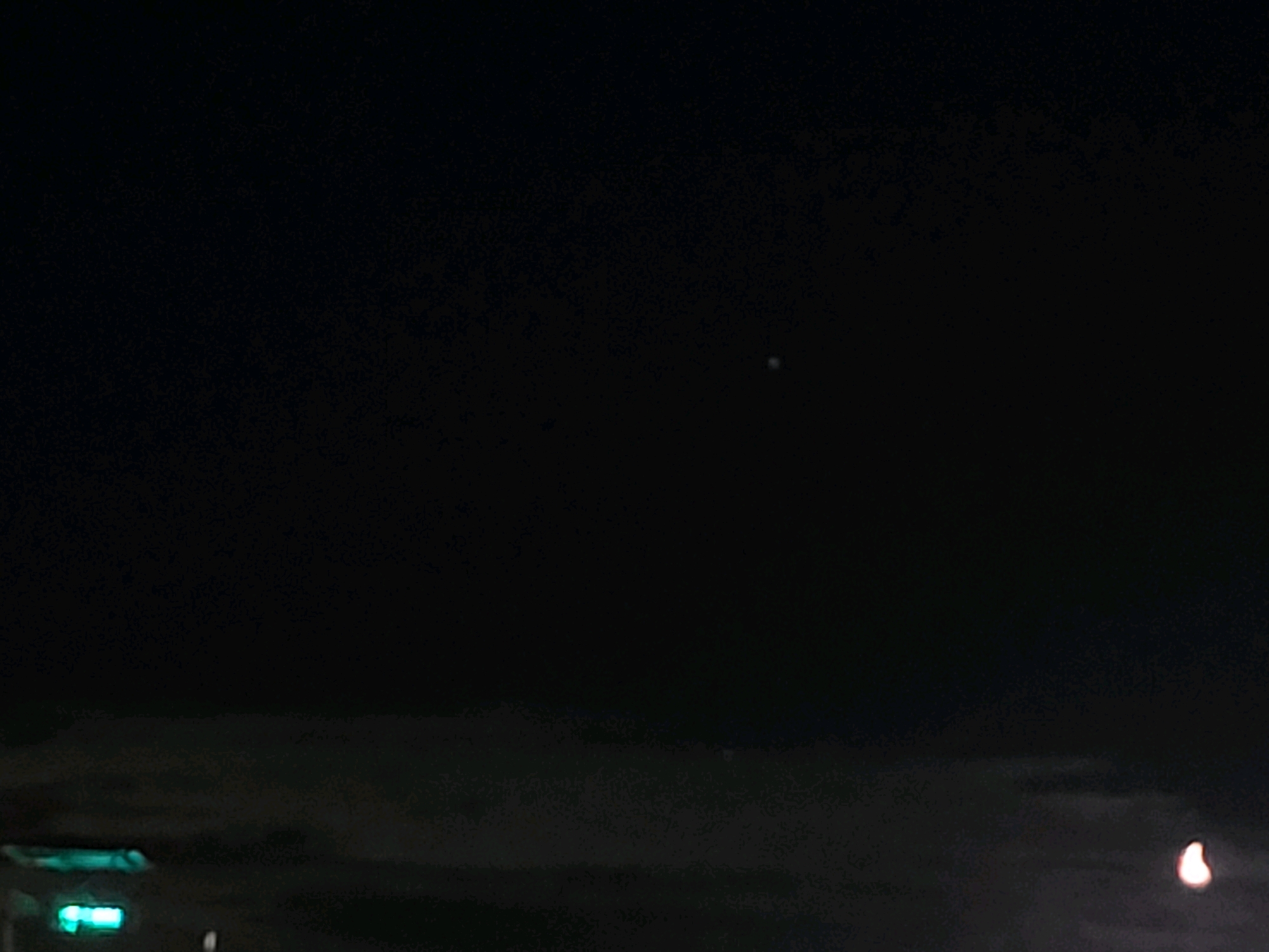 The image depicts a dark, possibly nighttime, urban or semi-urban environment. In the lower left, there are some indistinct green lights, possibly from a traffic signal or illuminated signage. The sky is predominantly black, with some very faint, grainy texture suggesting clouds or atmospheric conditions. Slightly above the center of the image, there is a small, bright, point-like object, likely a star or a distant light source. In the lower right, a blurry, light-colored object, possibly a distant light or a celestial body, is visible. The location is identified as Ozolnieki, Latvia.