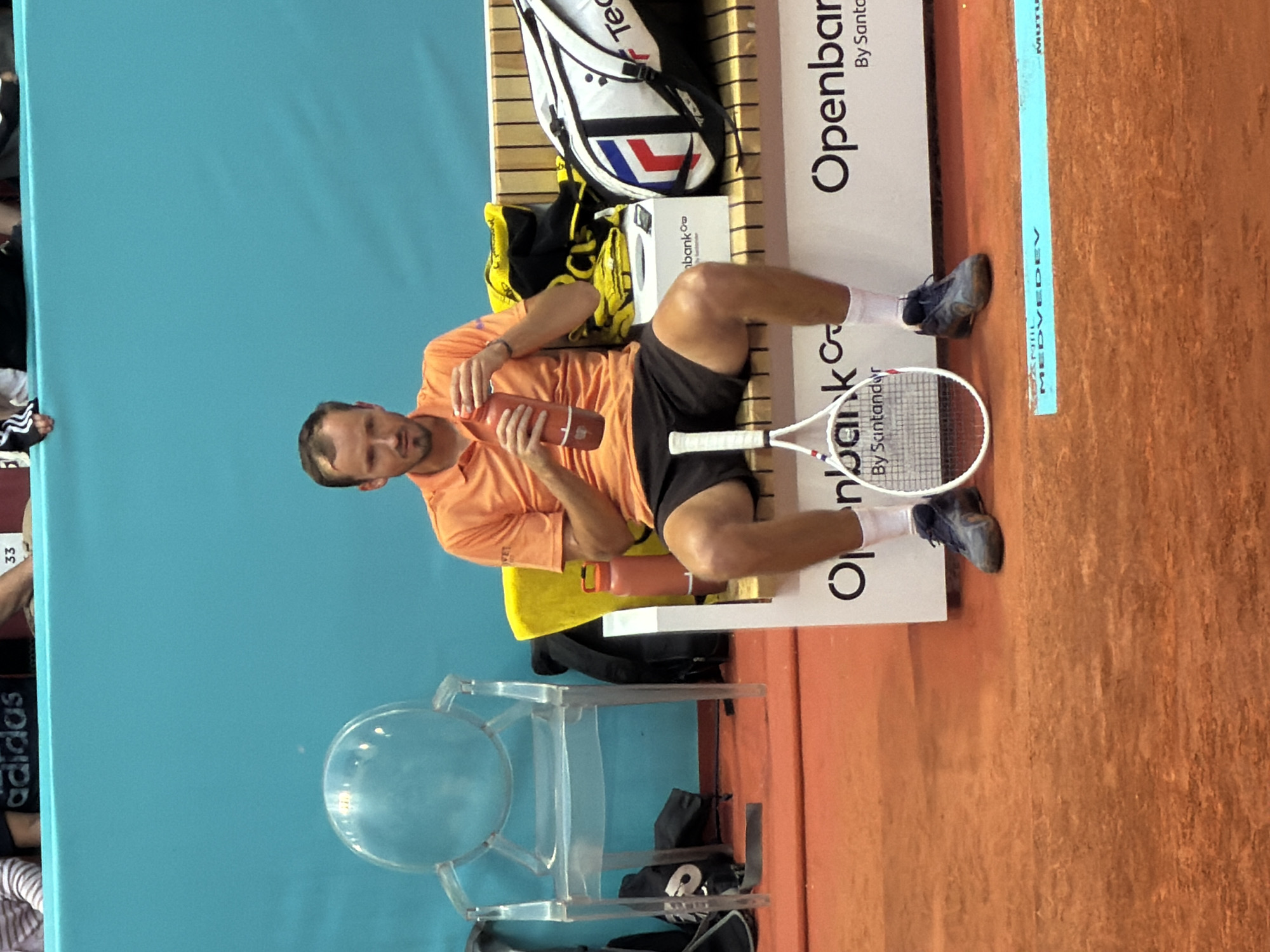 The image shows a man sitting on a bench during what appears to be a tennis match. He is wearing an orange shirt and black shorts and is holding a water bottle. A tennis racquet is resting on the bench next to him. Behind him is a light blue wall and a transparent chair. To his side is a sign for the Openbank Santander, indicating sponsorship of the event. A blue stripe on the ground has the lettering "Daniil Medvedev" on it. The surface on which the bench rests is reddish-orange, suggesting a clay tennis court. Given the event sponsorship and the surface, the scene is likely taking place at the Mutua Madrid Open tennis tournament in Madrid, Spain.
