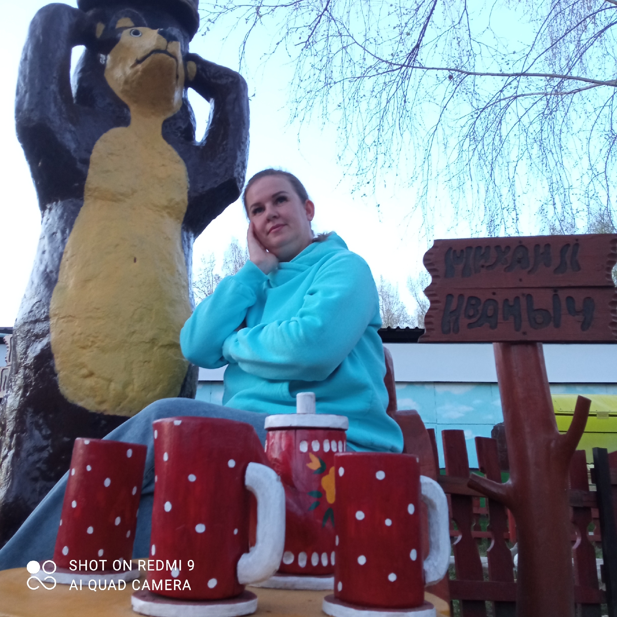 The image shows a woman sitting at a wooden table with decorative red mugs and a teapot in what appears to be an outdoor setting. A sign with Cyrillic text is visible in the background. Based on the sign reading "Mikhail Ivanych," along with other visual cues, the location is likely a children's playground or recreational area in the city of Chaikovsky, Russia. A statue of a bear stands beside the woman. She is wearing a light blue hoodie and jeans. She is leaning on her hand with her arms crossed. The sky is visible through bare tree branches.