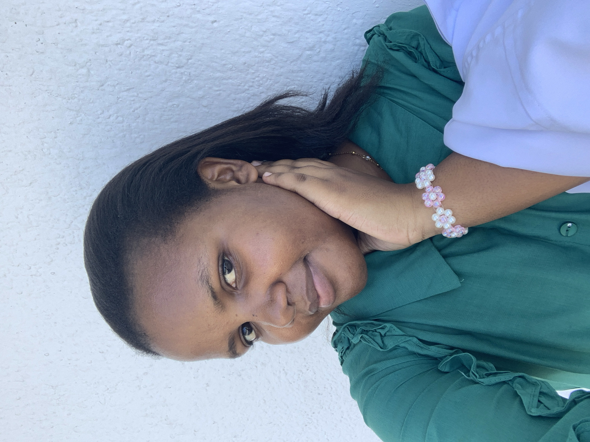A woman is pictured indoors, smiling. She wears a green blouse and a pink and white flower bracelet. Her left hand is touching her face. Part of a white top is visible in the upper right corner. The background behind the woman is a textured white wall. There is no information to specify that the image was taken in the city of Dar es-Salaam, Tanzania.