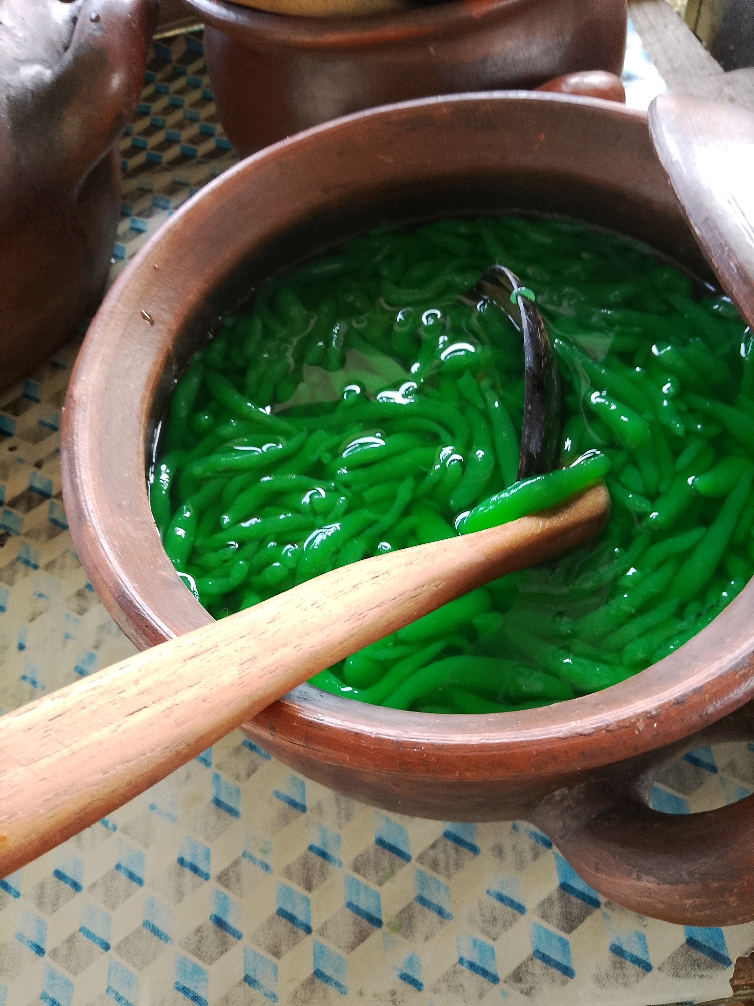 A close-up shot shows a traditional Indonesian dessert, likely Cendol, served in a rustic brown clay pot. The dessert consists of vibrant green, jelly-like strands submerged in a clear liquid, possibly coconut milk or syrup, with small white highlights suggesting the presence of ice or light reflection. A wooden spoon rests on the rim of the pot, its end dipping into the dessert, indicating it's ready to be served. The pot itself is positioned on a patterned surface with a geometric design of alternating blue and grey cubes against a white background. In the background, another similar clay pot is visible. The overall impression is one of a simple, refreshing, and authentic culinary presentation. The image is taken in Malangsari, Indonesia.