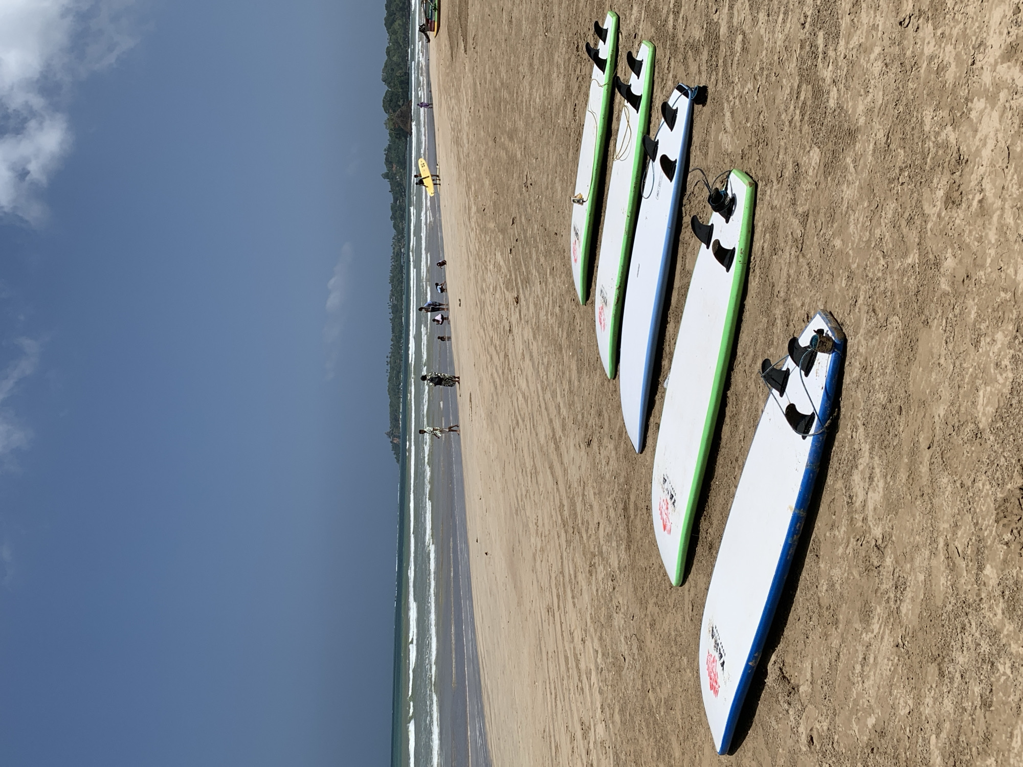 The image captures a beach scene in Weligama, Sri Lanka, with multiple surfboards lined up on the sandy shore in the foreground. The surfboards are predominantly white with colored trim, and some have fins visible. In the middle ground, the ocean displays moderate waves, and a few individuals are visible walking along the beach and in the water. Further in the background, a lush green coastline with scattered trees and structures suggests the town of Weligama. The sky is partly cloudy, with a mix of blue sky and white clouds. The overall impression is a tranquil beach day with opportunities for surfing.