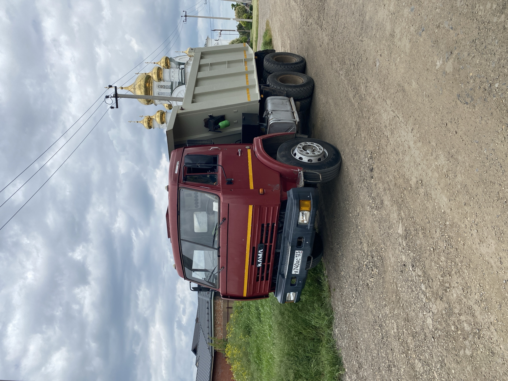 A red KAMAZ truck with license plate A740AC123 is parked on a dirt road in Bryukhovetskaya, Russia. The truck is facing the camera. A church with golden domes is visible in the background. The sky is cloudy. A building with a brick facade is visible in the lower left corner of the image. Power lines are also visible in the background.