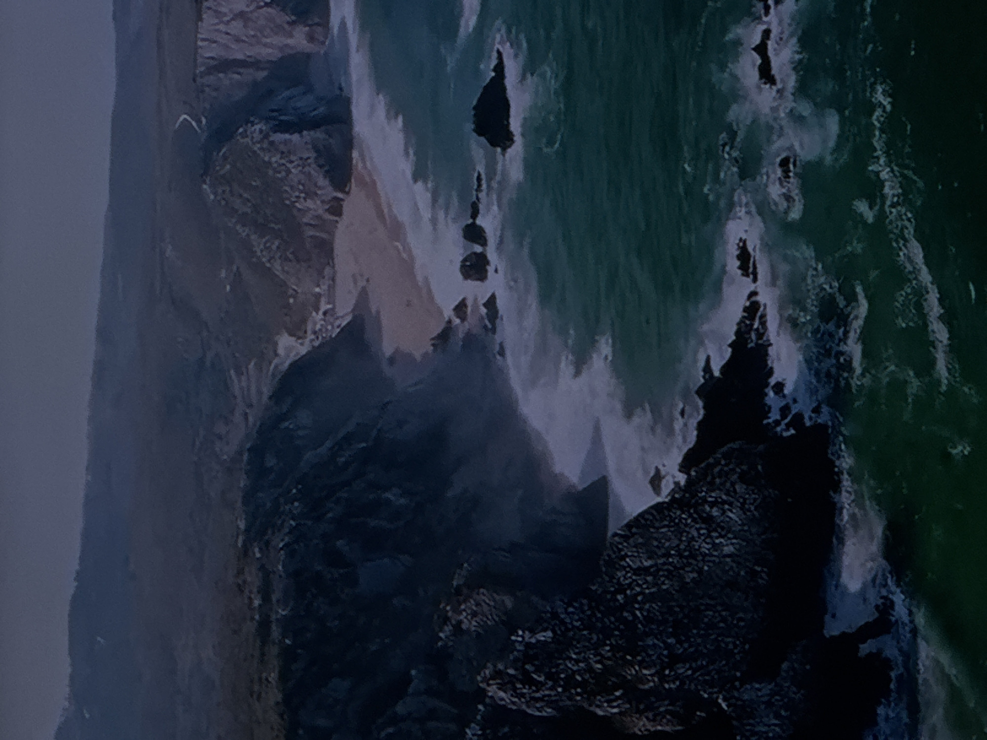 This is an aerial, high-angle, long shot of a rocky coastline with turquoise ocean waves crashing against the shore. The sea is turbulent, with white foam and spray visible. Several dark, jagged rocks jut out of the water near the coastline. A small sandy beach is nestled between the cliffs. The cliffs themselves are steep and layered, with dark shadows. In the background, there are rolling hills and a faint, winding road. The sky is a dusky, overcast blue. The scene is devoid of human presence and appears to be a natural landscape. The location is not within the city of Sinyavino, Russia.