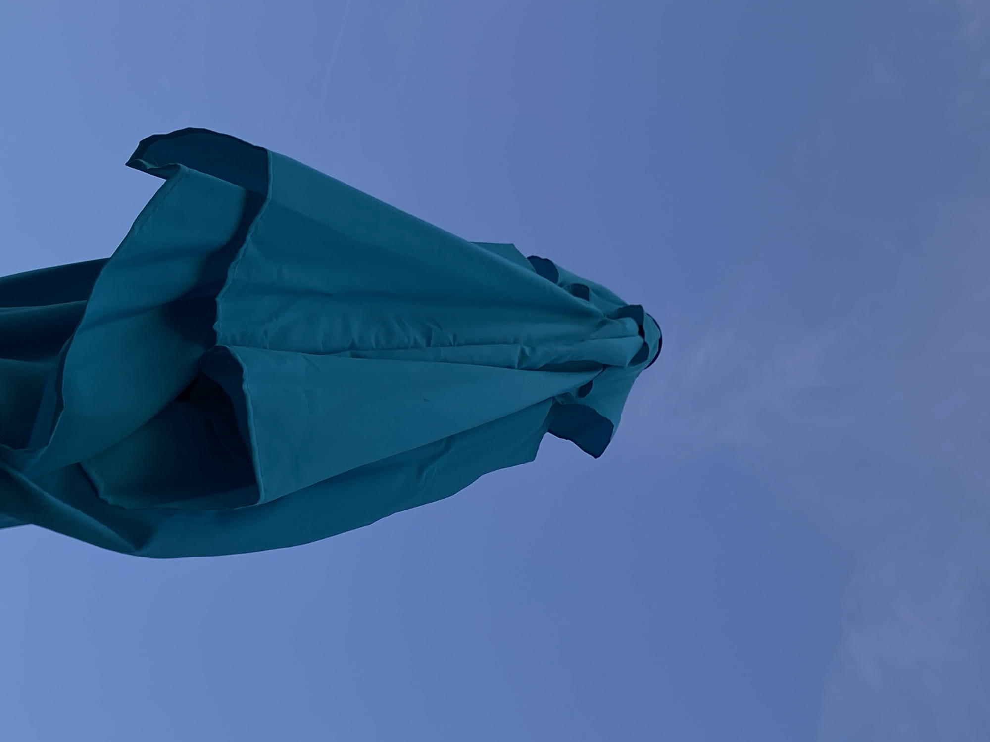 A close-up, low-angle shot shows a closed teal-colored patio umbrella against a deep blue sky with wispy white clouds. The umbrella is the primary subject, occupying the majority of the frame. Its fabric is gathered and bunched, creating folds and shadows that give it a sculptural appearance. The umbrella's top, where the fabric meets the pole, is visible, revealing a dark rim. The sky is a vast expanse, with the blue deepening towards the top of the image. The lighting suggests it is either early morning or late afternoon, as the sky is not bright white and the umbrella is not brightly illuminated. The image was taken in the city of Arandjelovac Municipality, Serbia.