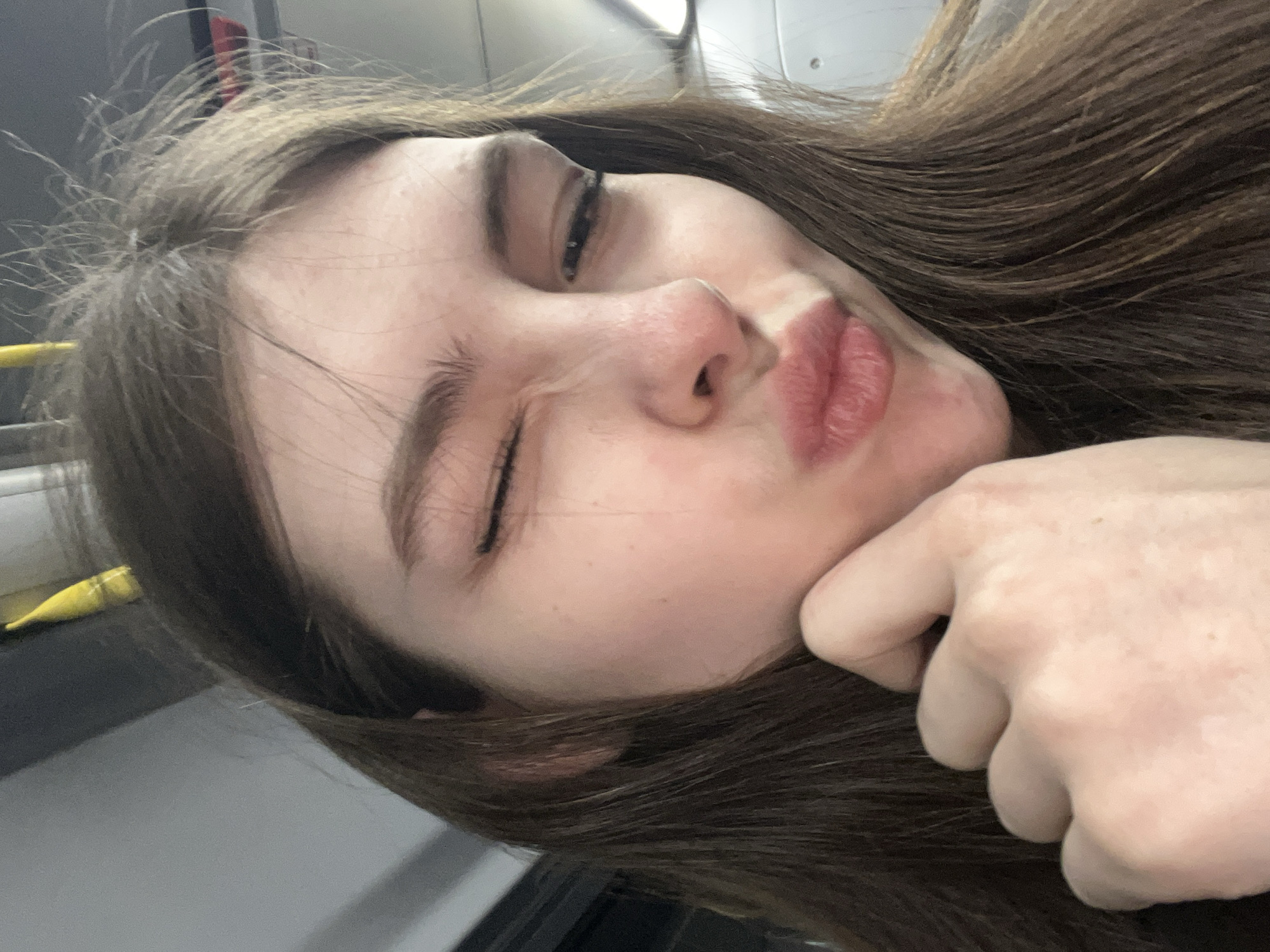 A young woman with long brown hair is holding her face with her right hand. Her right eye is almost closed, and she is pouting. She is seated on a bus. The location is unknown.