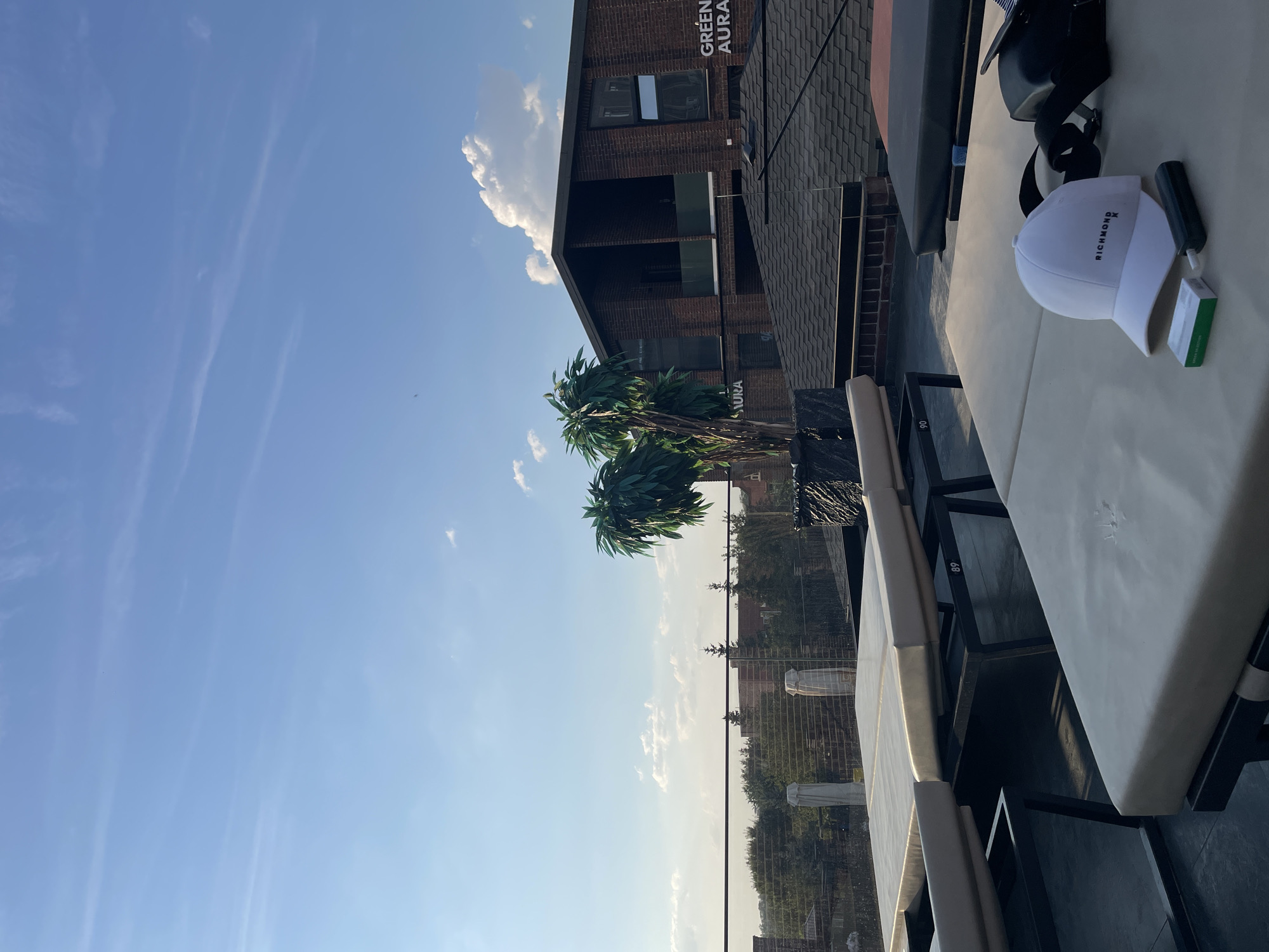This is a rooftop scene with lounge chairs and a building in the background, set against a clear blue sky with some clouds. The building appears to be made of brick and has large windows. There are artificial palm trees on the rooftop. In the foreground, there are lounge chairs and a white baseball cap with the word "RICHMOND" and a logo on it. Next to the cap are a small green and white box and a dark cylindrical object. The location is specified as Leninavan, Russia.