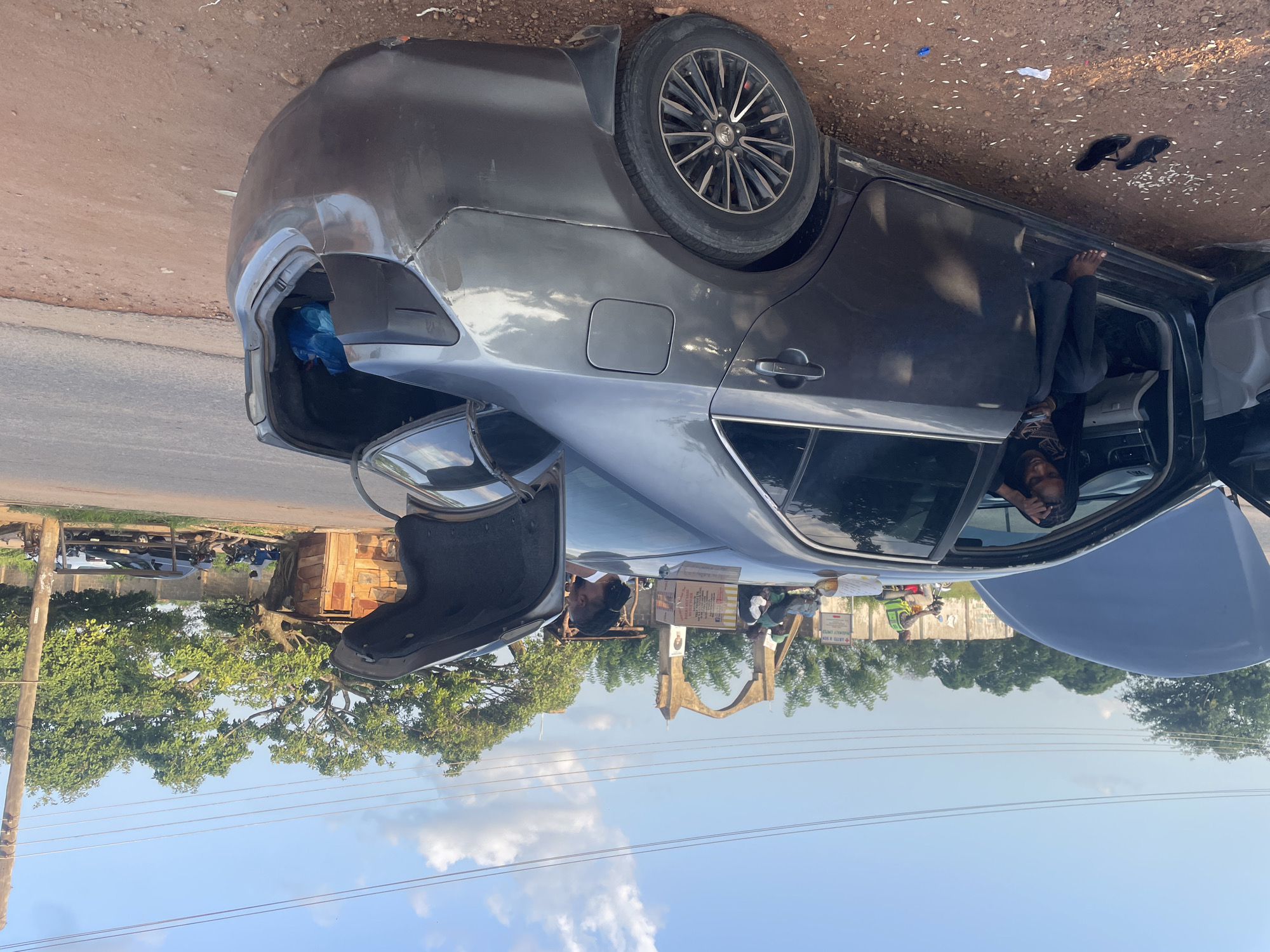 An eye-level, medium shot captures a gray sedan with its hood and trunk open, parked on a dirt shoulder next to a paved road. The car appears to be in a residential or semi-rural area of Kabba, Nigeria, with a wooden structure and large trees visible in the background. A young woman with braided hair sits in the driver's seat, looking at her phone. Another woman, partially visible, stands near the open trunk, which contains a blue bag. Scattered white grains and a pair of black flip-flops lie on the ground beside the open car door. In the distance, a motorcyclist in a high-visibility vest rides past a building with an arched entrance, suggesting a bustling environment despite the seemingly stalled vehicle. The sky is partly cloudy, and the sunlight casts shadows, indicating it is either late morning or late afternoon.