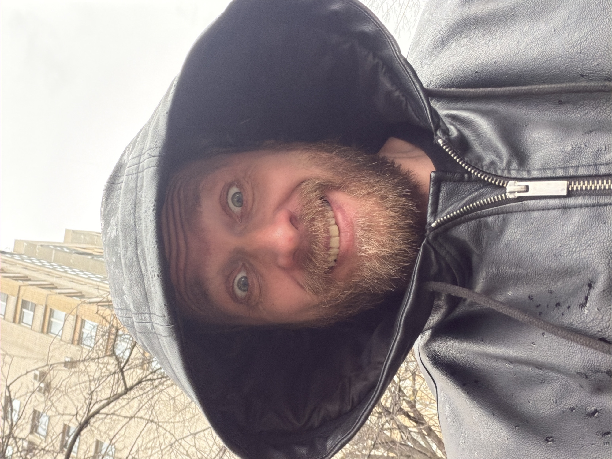A man with a beard smiles at the camera while wearing a black leather jacket with the hood up. He appears to be outside, with leafless trees and a light-colored building with windows visible in the background. Given the architectural style, the setting is potentially Saint Petersburg, Russia. The jacket is slightly wet. The man's expression is cheerful.