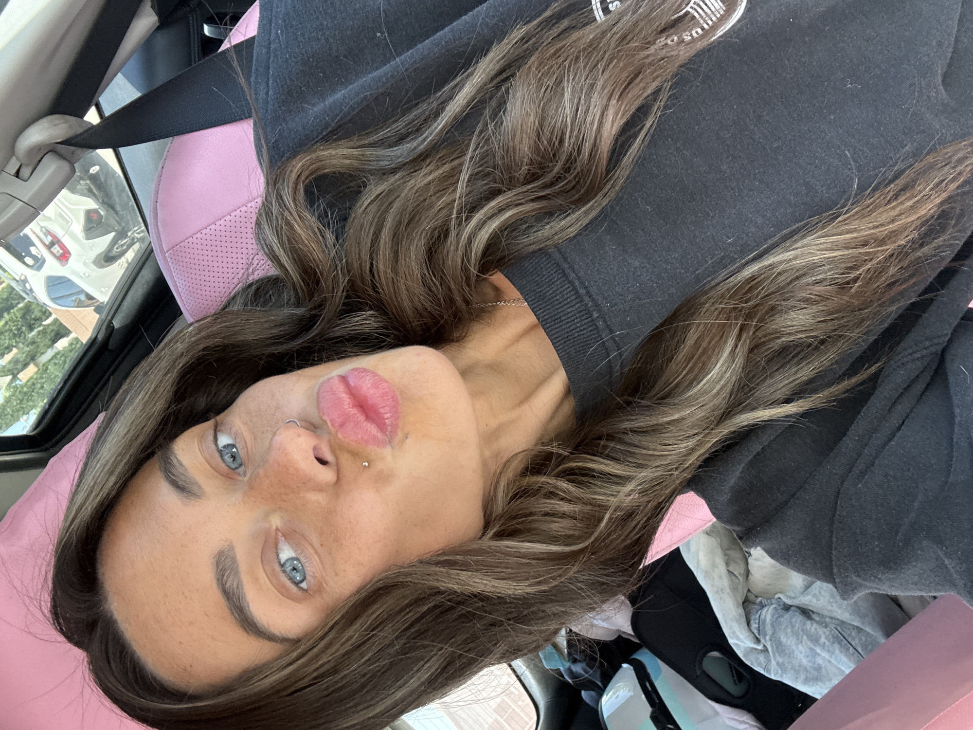 A selfie-style portrait captures a young woman with long, wavy brown hair featuring lighter highlights, positioned horizontally in a car. She has striking blue eyes, well-defined dark eyebrows, and is making a "kiss" face with her full, pink lips. Her face features several small silver piercings: a ring in her left nostril, a stud above her upper lip, and another below her lower lip. She wears a dark grey or black long-sleeved sweatshirt, with an indistinct white logo or text partially visible on the upper left chest area. A thin silver necklace is also visible around her neck.
The setting is inside a car, with a bright pink, perforated leather car seat or seat cover behind her head and shoulders. A black seatbelt runs diagonally across the upper left of the frame. Through the car window, an outdoor environment is visible, featuring other vehicles, including a white SUV and a darker car, along with some distant greenery, suggesting a parking lot or street scene during daylight. Various folded items of clothing and bags are casually placed on what appears to be an adjacent seat or the car floor. The woman's pose conveys a playful or confident mood, captured casually in Bellflower, United States of America.
