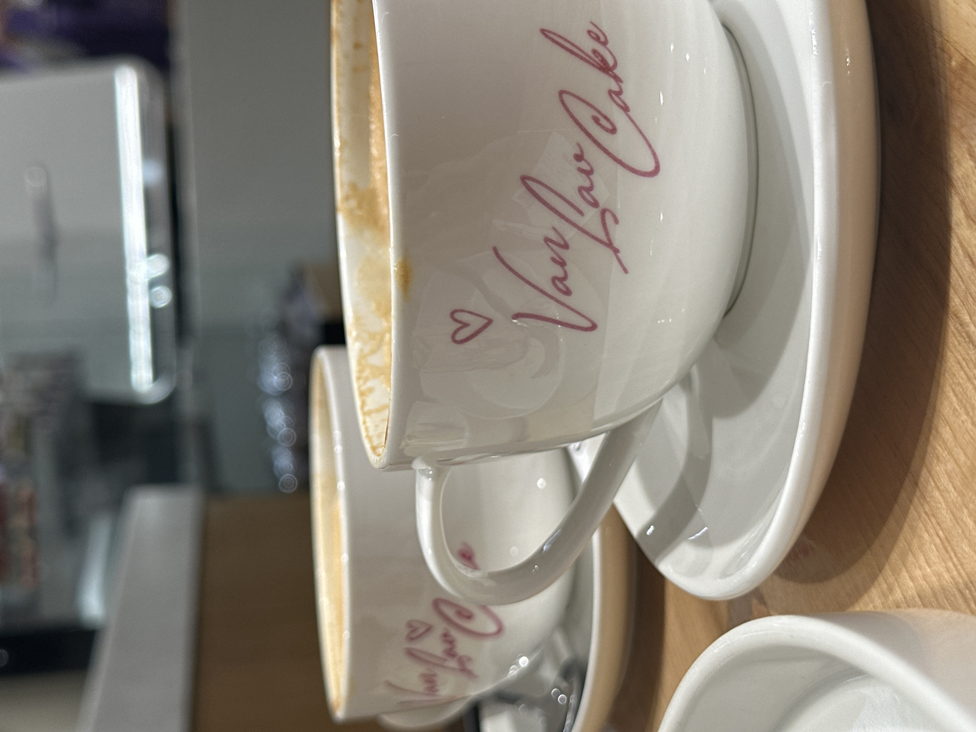 The image shows several white ceramic cups and saucers stacked on what appears to be a wooden surface. The cups have the words "Van Lav Cake" written on them in pink script, along with a small heart symbol. The cups contain remnants of a beverage, possibly coffee or tea. In the blurred background, there's a hint of a machine, possibly a coffee maker or dispenser, and other unidentifiable objects. There is no indication in the image of its location in Rostov-on-Don, Russia.