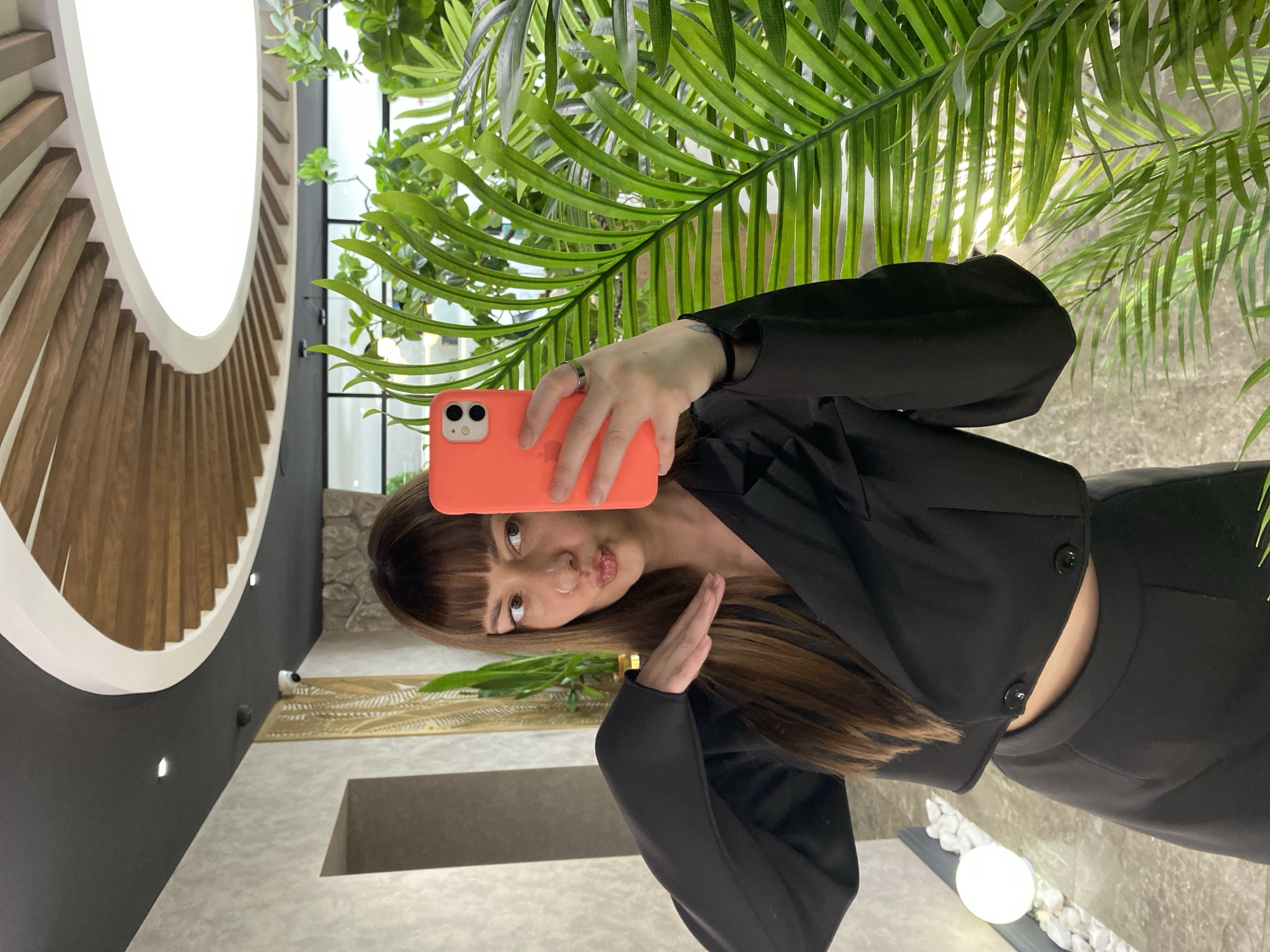 A young woman with brown hair and bangs takes a selfie with an orange smartphone. She is wearing a black suit with button details and a ring on her finger. The background features lush green plants and a modern interior with a decorative lighting fixture. The location is in an indoor area within the city of Votkinsk, Russia.