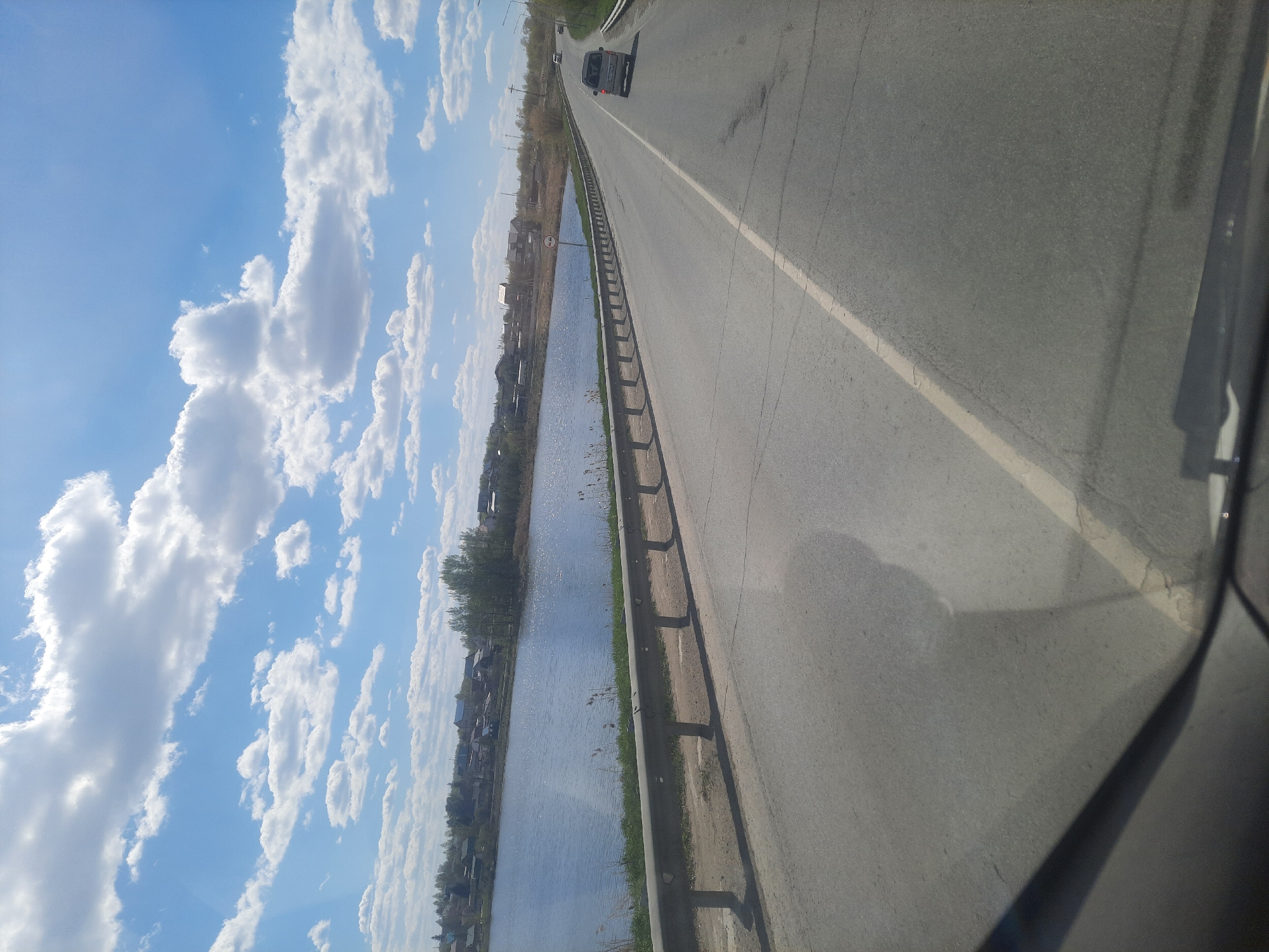 The image depicts a view from a vehicle traveling on a road in Научный Городок, Russia. The road runs parallel to a body of water with vegetation and buildings visible on the opposite shore. A guardrail separates the road from the water. The sky is blue with scattered clouds. A vehicle is ahead on the road.