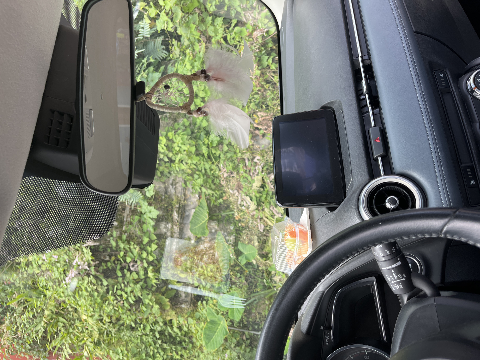 The image presents an interior view of a vehicle, looking out through the windshield into a lush, green natural environment, consistent with the tropical landscape of Cuetzalan, Mexico. The perspective is from the driver's side, showing parts of the dashboard, steering wheel, and the rearview mirror.
In the upper center, a rearview mirror is prominent, from which a decorative dream catcher with a woven center and light-colored feathers hangs. To the right on the dashboard, a small rectangular digital screen, likely a GPS unit or dash camera, is mounted. Further right, a hazard light button with a red triangle symbol is visible, alongside a circular air vent.
The steering wheel is partially seen in the lower right, with a multi-function stalk extending from behind it. Text on this stalk includes "OFF," "INT," "ON," "AUTO," "LO," "HI," "MIST," "PULL," and "WASH," indicating wiper controls. A button nearby reads "TRIP."
On the dashboard, near the windshield, there's a small clear plastic container, possibly holding fruit, and reflections on the glass show what appears to be a fork and other interior elements.
Outside the vehicle, the scene is dominated by dense, vibrant green foliage, including various trees and plants, suggesting a rich, natural setting. The bright, even lighting indicates it is daytime. The car appears to be stationary or slowly moving through a scenic, verdant landscape. No people are visible.