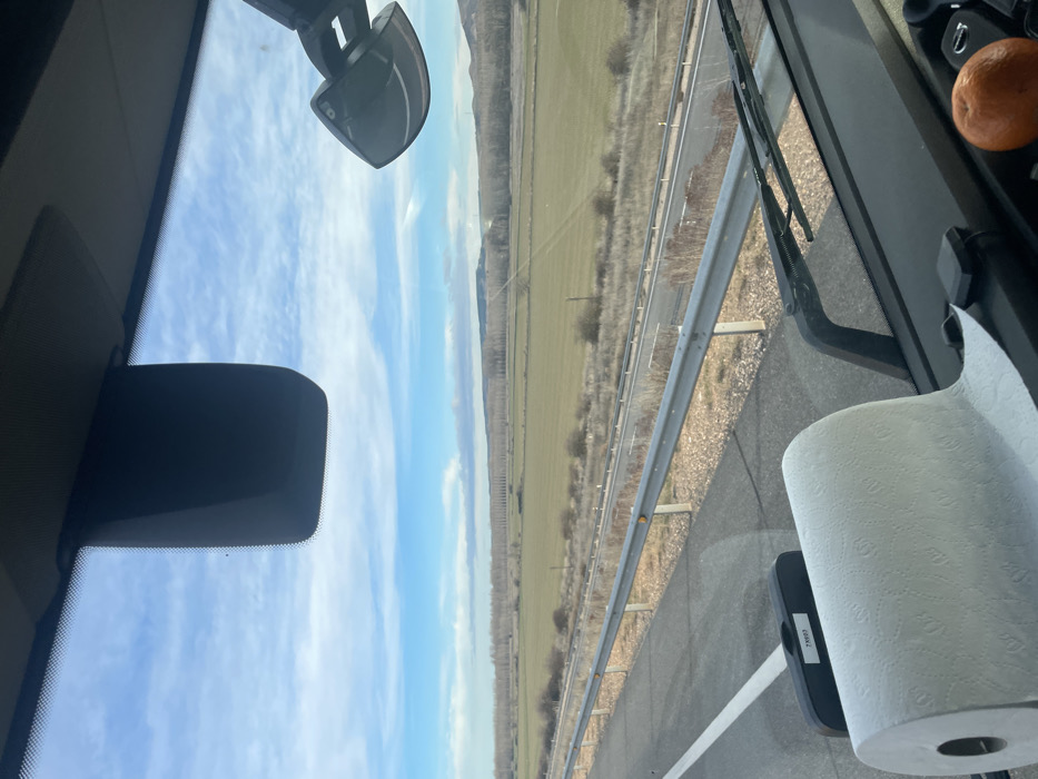 From inside a vehicle, a view shows a road alongside a field with a line of trees. The sky has blue and white clouds. The vehicle's interior features a rearview mirror, a sun visor, a roll of paper towels, an orange, and windshield wipers. The image is taken through the windshield.