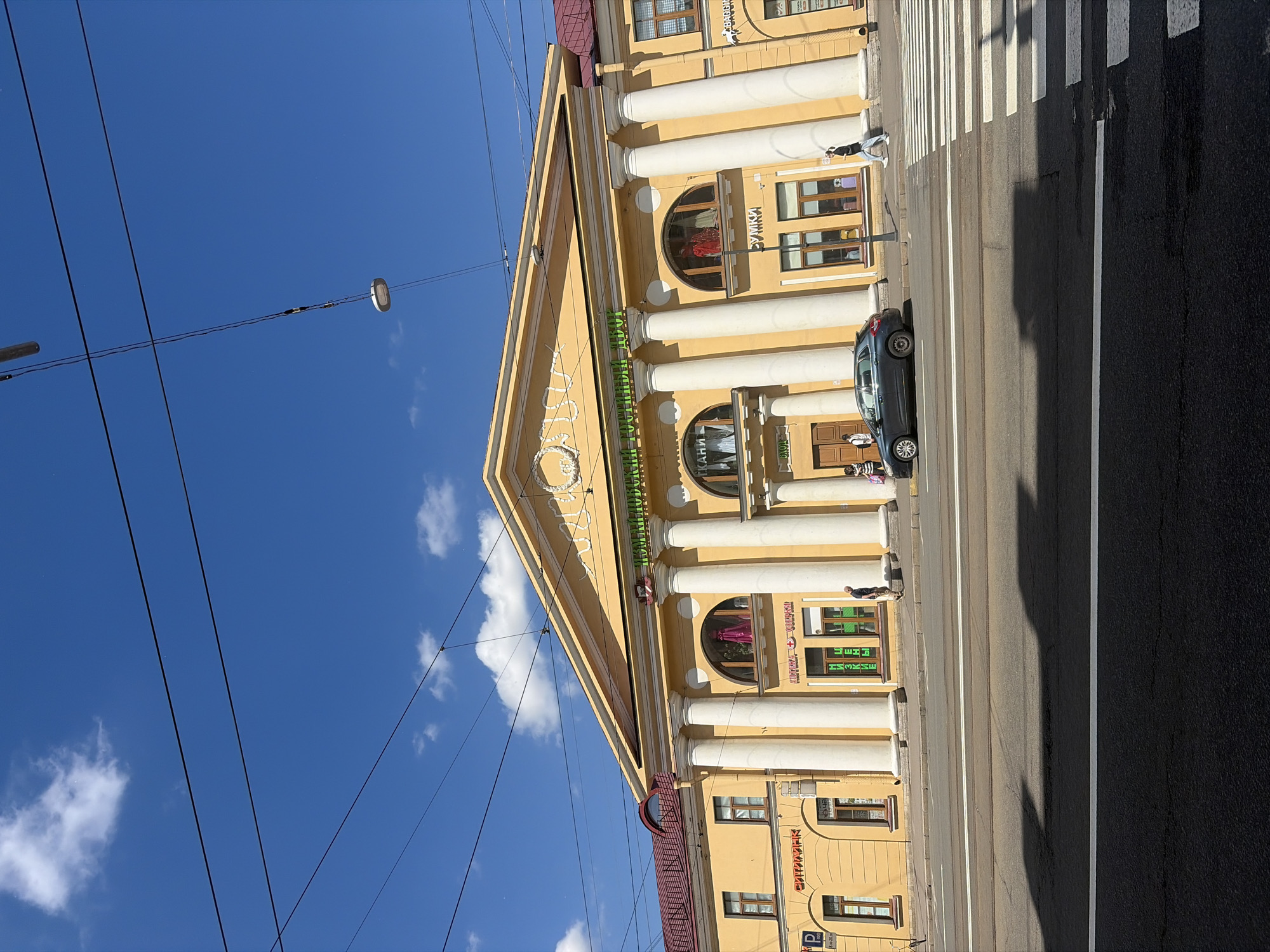 The image shows a sunny day in front of a yellow building in Saint Petersburg, Russia, featuring a pediment supported by large columns. The building has arched windows and a sign on the top facade that reads "Гостиный Двор" (Gostiny Dvor). A dark-colored hatchback car is parked on the street in front of a pedestrian crossing. Several people are visible walking on the sidewalk. The sky is blue with some scattered white clouds, and overhead, power lines are strung across the frame. The building's architecture suggests it is a historic landmark in the city.