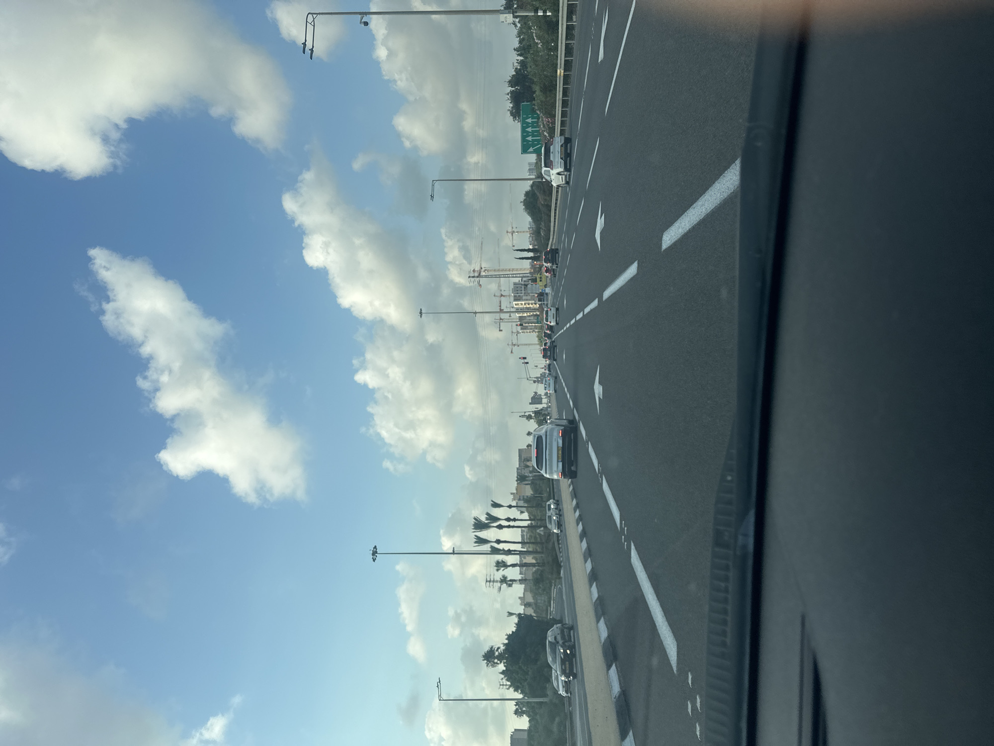 The image shows a view from the perspective of a vehicle driving on a multi-lane highway, likely in or near the city of Acre, Israel, given the context. The sky is blue with scattered white clouds. Along the right side of the highway, there is dense green foliage and trees, with some flowering bushes. Further in the background, there are buildings under construction with several large cranes visible. The highway itself has clear white lane markings, indicating multiple lanes of traffic. Several vehicles are on the road, including a white SUV in the right lane, a black SUV in the left lane, and other cars further ahead. There are streetlights and utility poles lining the highway. The overall impression is of a modern urban or suburban environment with ongoing development.