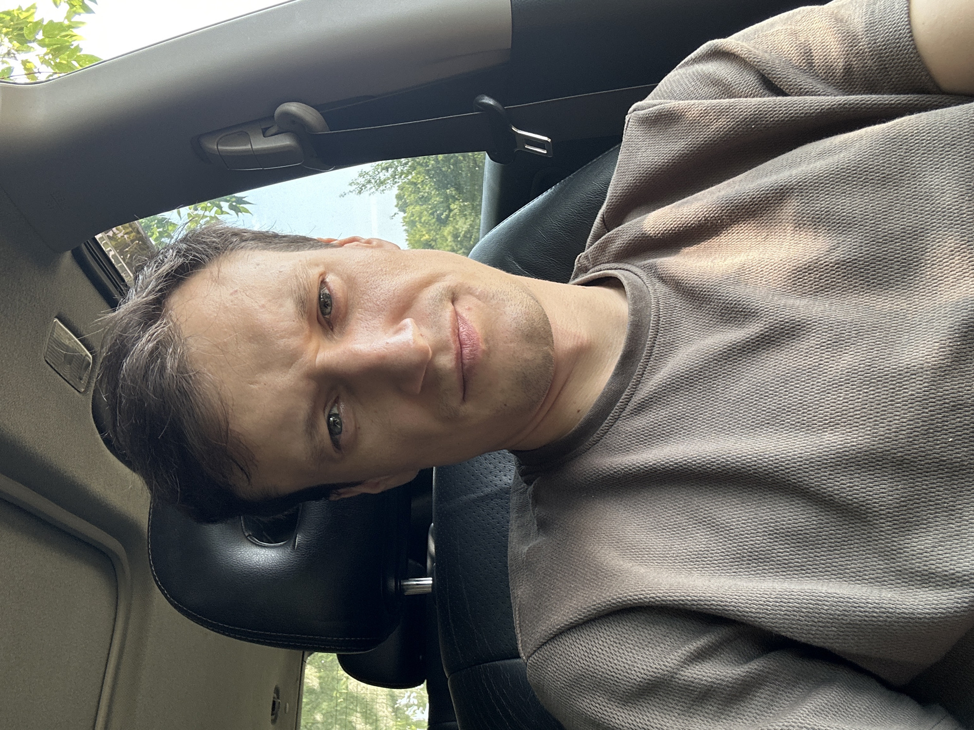 A close-up, selfie-style shot features a white man with short brown hair and green eyes. He is seated in the driver's seat of a car, with the interior visible, including the headrest of the passenger seat and a partial view of the car's roof with a sunroof. The man is wearing a tan, textured t-shirt. Sunlight is streaming into the car, casting shadows and highlights on his face and shirt. Through the car windows, trees and a bright sky are visible, suggesting an outdoor, possibly rural, location. The man's expression is neutral, with a slight furrow in his brow. The image appears to have been taken during daylight hours. The location is specified as Makiv, Ukraine.