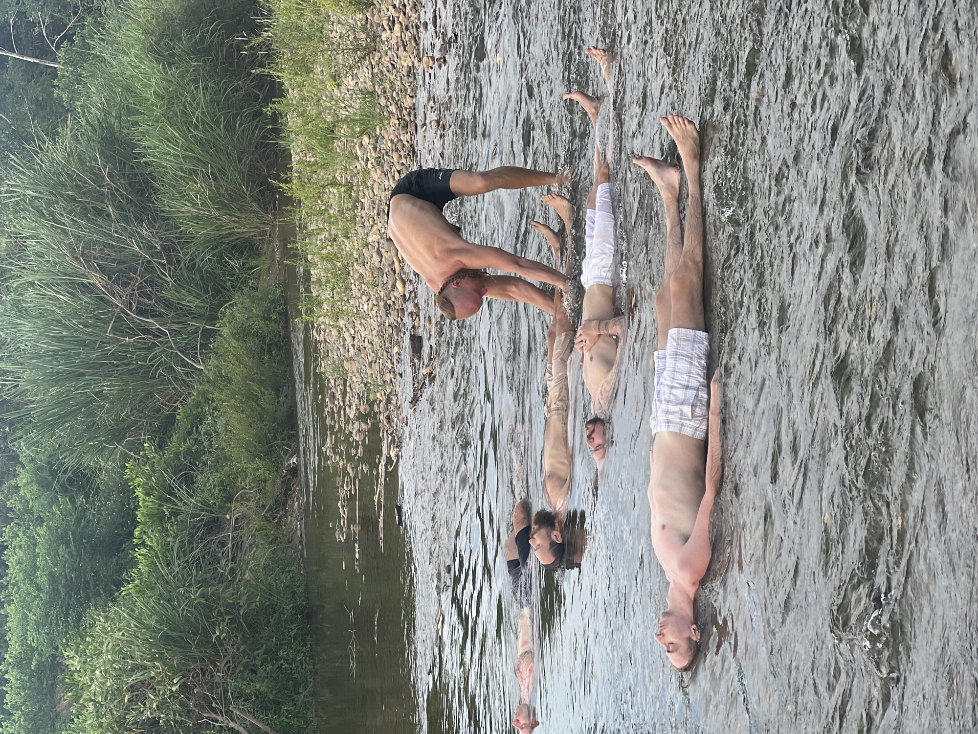 A group of men are floating on their backs in a shallow river, with a single man standing and interacting with them. The riverbed is visible, covered in stones and pebbles. Lush green vegetation, including tall reeds and trees, lines the riverbank in the background. The water is clear with gentle ripples. The standing man is wearing black shorts, and the floating men are wearing various shorts or swim trunks. The overall scene appears to be a therapeutic or wellness activity taking place in a natural outdoor setting. This scene is not specific to Đà Nẵng, Vietnam.