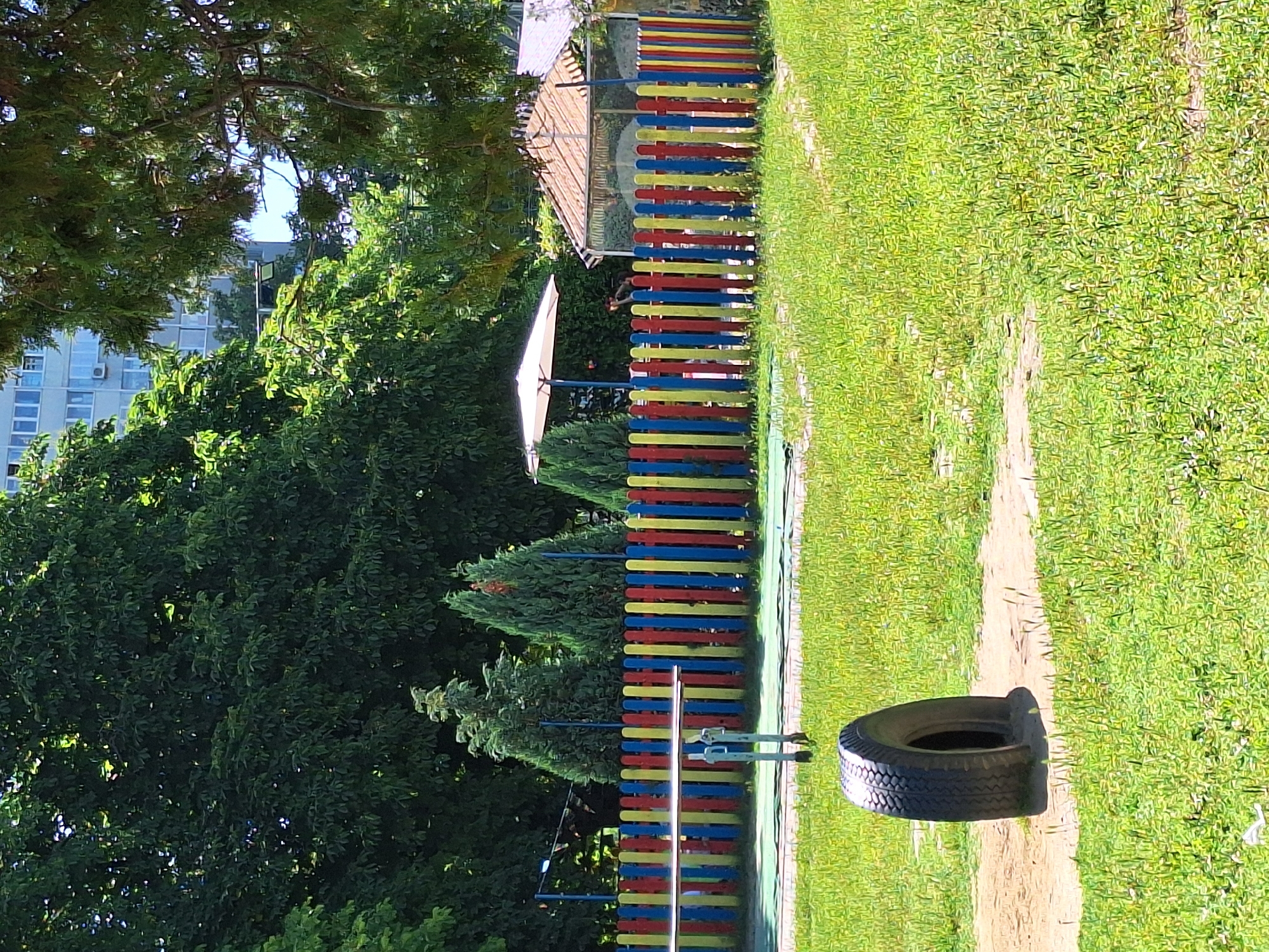 The image depicts an outdoor scene in Varna, Bulgaria, likely a park or playground. The primary subjects are a vibrantly colored fence, lush green grass, and trees. The fence is made of vertical planks painted in alternating red, yellow, and blue stripes. Behind the fence, there's a glimpse of a structure with a light-colored roof and a white umbrella.
To the right of the fence, a dirt path winds through the grassy area, and a single tire lies on the path. In the background, dense green trees dominate the left side of the frame, with a modern building visible through a gap in the foliage. The overall scene suggests a sunny day, given the bright illumination and clear skies hinted at through the trees. The presence of a playground fence and tire suggests the area is designed for recreation, possibly for children. No people are visible in the image, and no text is present. The orientation of the image is skewed, appearing rotated.