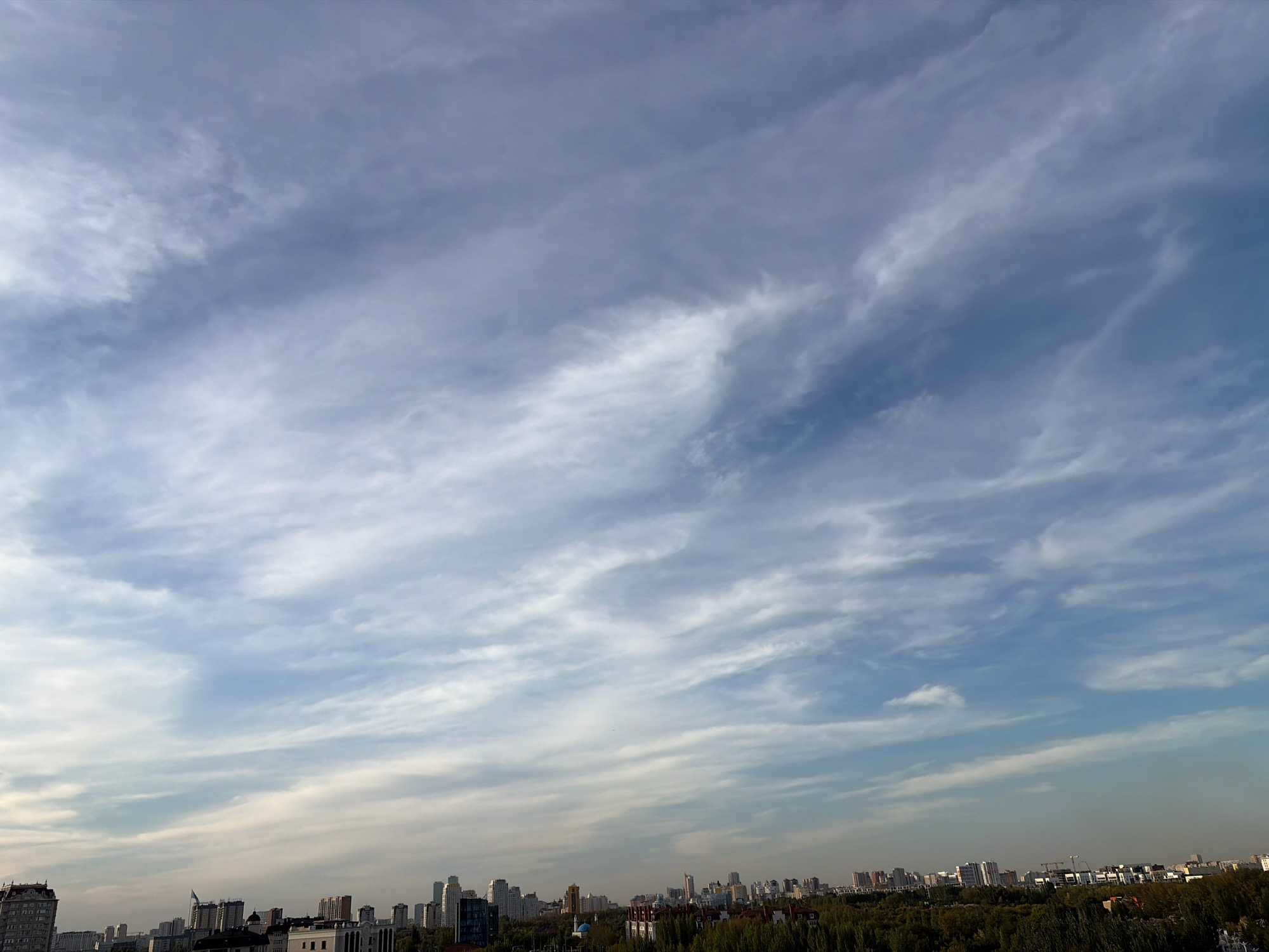 The image displays a panoramic view of the Astana, Kazakhstan skyline under a vast, partly cloudy sky. The clouds are wispy and stretched across the blue expanse, creating a dynamic texture. In the foreground, a dense line of trees forms a green band separating the sky from the urban landscape. The city itself is characterized by a mix of modern high-rise buildings and smaller structures, interspersed with cranes indicating ongoing construction. The architecture suggests a contemporary urban environment. No people are visible in the frame, and the scene is static, depicting a typical city view from a high vantage point.