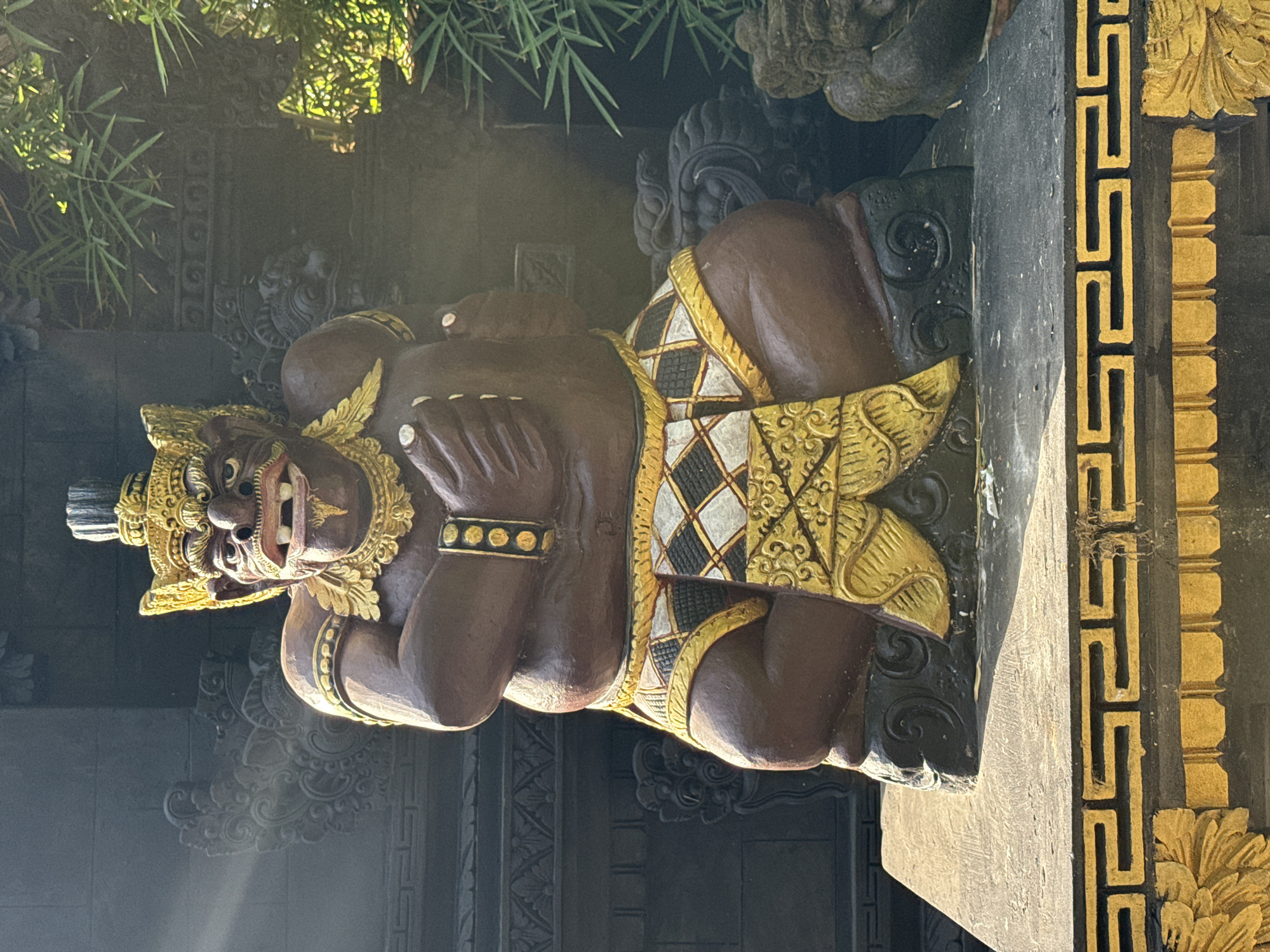 The image displays a seated, robust statue of a guardian figure, typically found in Balinese culture, crafted from a dark brown material with gold and white accents. The figure has a broad, expressive face with prominent fangs or teeth, a wide mouth, and large eyes under a golden, elaborate crown. A golden necklace and armbands adorn its upper body. A patterned skirt or sarong, featuring black and white diamond checks with gold borders and a lower gold scrollwork design, covers its lower body. The figure's hands are positioned in front of its chest, holding small, white spherical objects.
The statue rests on a dark, carved circular base, which in turn sits on a rectangular, light-colored stone platform. This platform is edged with a repeating gold meander pattern and features additional gold floral carvings and vertical striations on its lower section.
The background on the left reveals a dark, intricately carved stone wall with traditional Balinese relief patterns, illuminated by diagonal rays of sunlight streaming from the upper left. On the right, dark stone structures are partially obscured by bright green, leafy tropical foliage, likely bamboo, also highlighted by sunlight. This scene is characteristic of a temple, traditional compound, or resort garden, commonly found throughout Kuta Selatan, Bali, Indonesia.