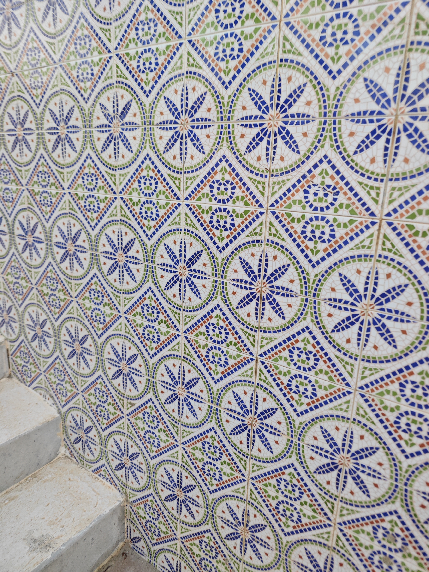 The image shows a wall covered in traditional Tunisian zellige tiles, featuring a geometric pattern in blue, green, and orange against a white background. The tiles are arranged in a repeating motif of starbursts enclosed within circles and diamonds. In the lower left corner, a concrete staircase is visible, suggesting the wall is part of an outdoor or transitional space. The image is likely taken in Mahdia, Tunisia, a city known for its historic medina and intricate tilework.