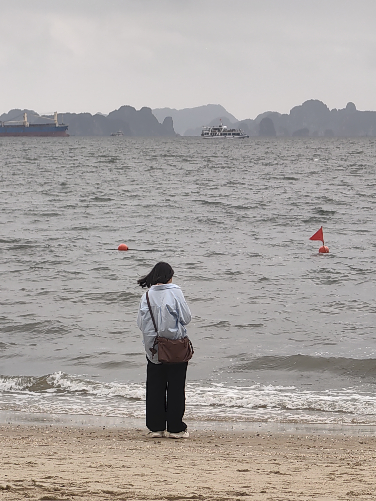 A person stands on a sandy beach facing the ocean in Bai Chay Ward, Vietnam. The person has dark hair and is wearing a light blue shirt, dark pants, and white shoes, with a brown bag slung over their shoulder. The ocean is choppy with small waves breaking on the shore. In the distance, there are islands and boats on the water. There is an orange buoy and a red buoy with a red flag floating in the sea. The sky is overcast.