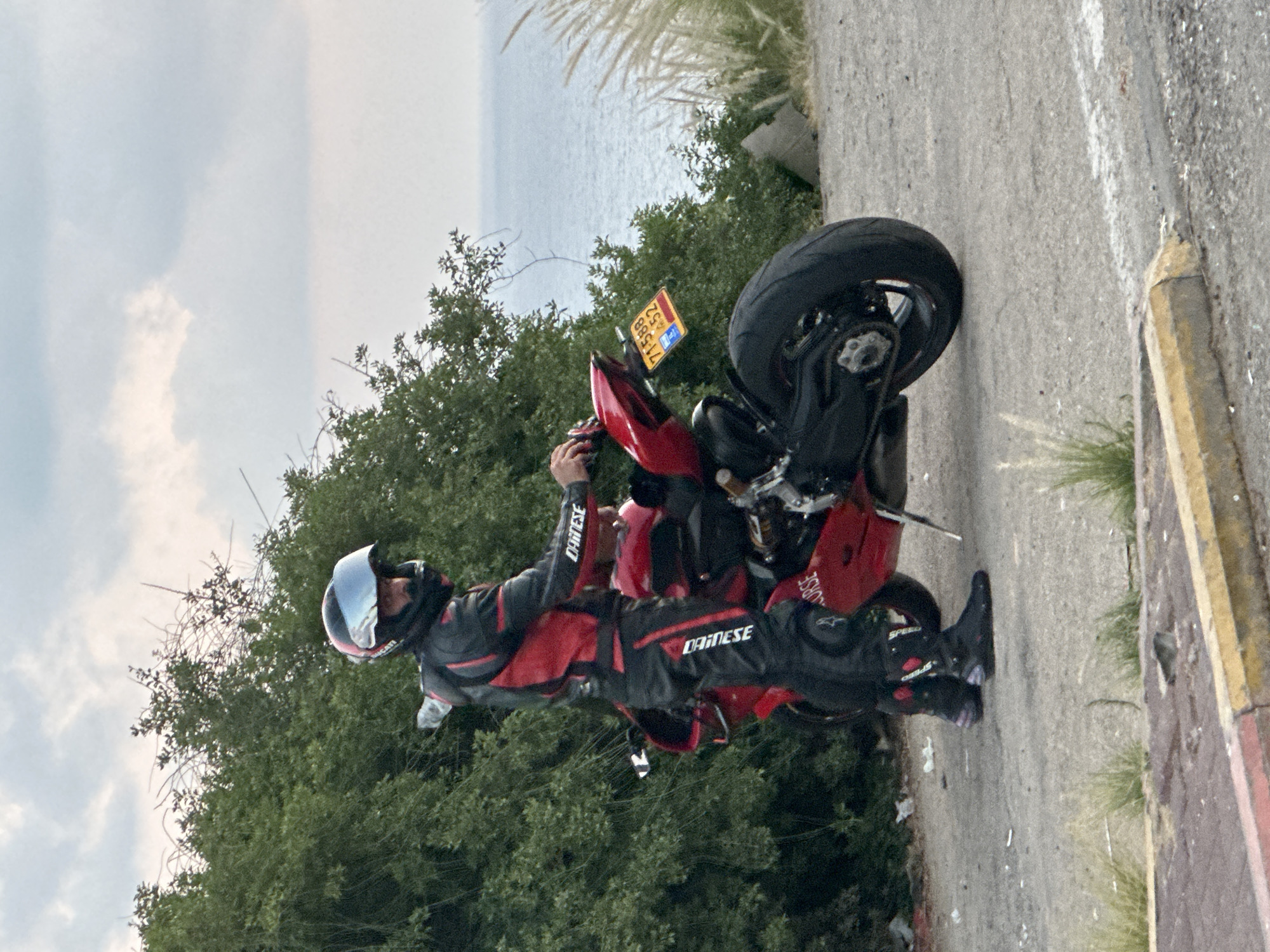 A motorcyclist wearing a red and black Dainese racing suit and helmet stands next to a red sport motorcycle. The motorcyclist is facing away from the camera and towards a body of water, possibly the sea. The motorcycle has a yellow license plate. The scene is set outdoors, with green foliage and the sea in the background. The ground is paved. The image is likely taken in a coastal area in the Mate Asher Regional Council, Israel, given the license plate and the vegetation.