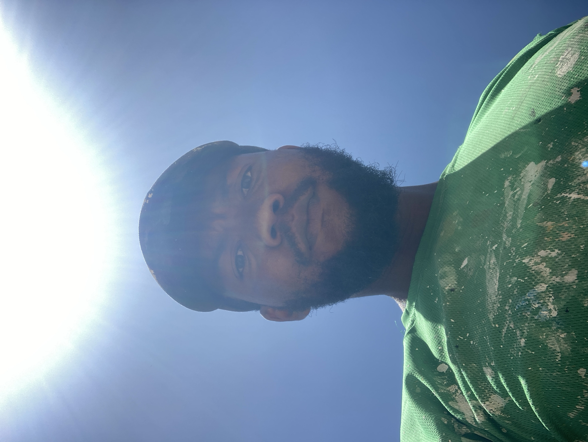 A man with dark skin, a full beard, and wearing a dark cap is looking down at the camera. He has on a green t-shirt with white paint stains. The sky is a clear blue with the sun shining brightly in the left portion of the frame. The location is not discernible from the given image.