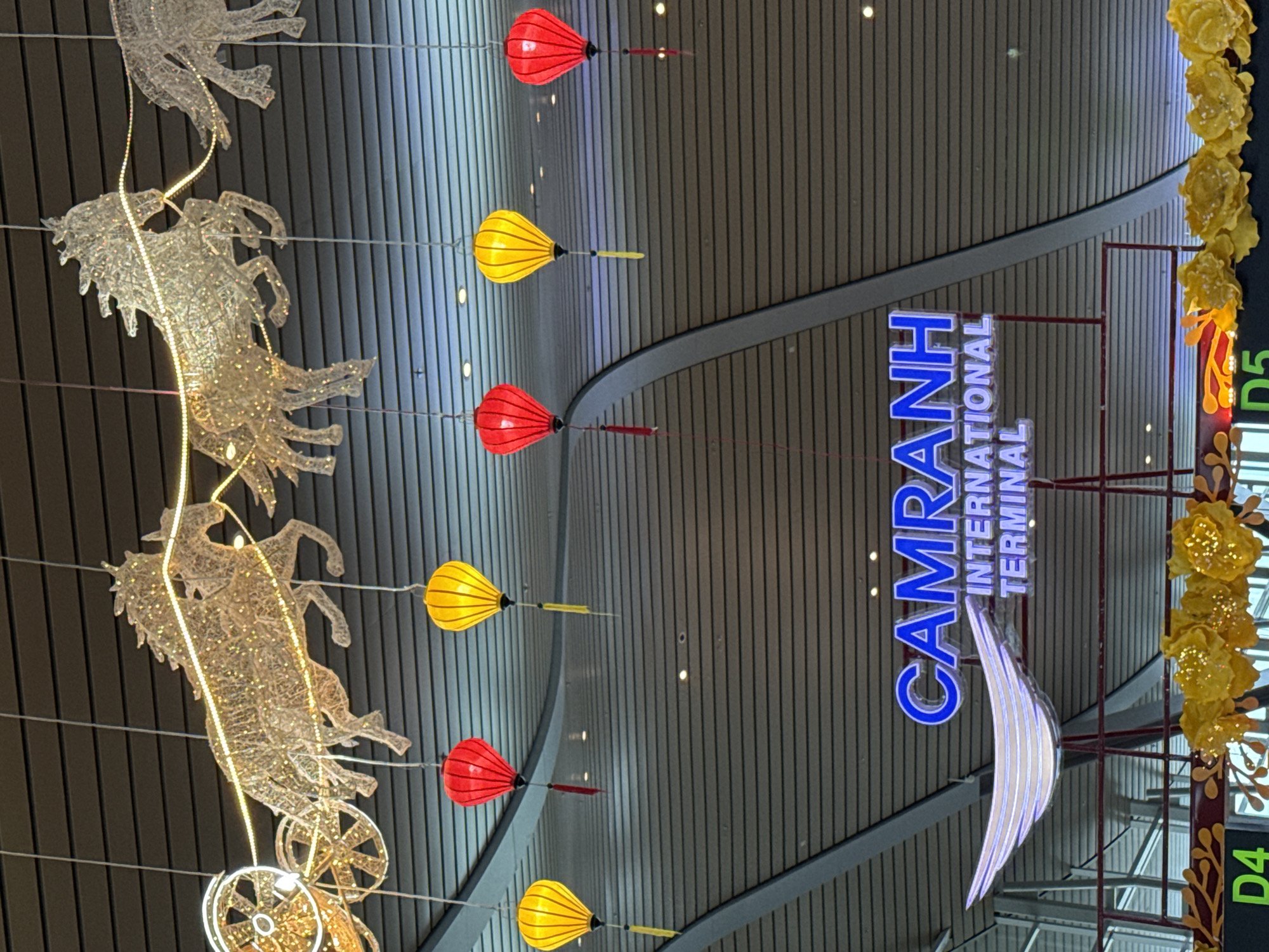 The image shows a decorative ceiling in a modern building, likely an airport terminal, with the name "CAMRANH INTERNATIONAL TERMINAL" prominently displayed in blue neon letters. Overhead, illuminated horse-drawn carriage figures made of wire and lights are suspended, creating a festive and dynamic visual. Below these, traditional Vietnamese lanterns in red and yellow hang at various heights, adding color and cultural flair. A stylized, glowing wing-like symbol is also visible near the signage. The ceiling itself is constructed of dark horizontal panels, with some recessed lighting visible. The lower part of the image shows part of a railing and some floral decorations, suggesting a celebratory or welcoming atmosphere. The overall impression is one of a well-decorated interior space, possibly for an event or a holiday.
