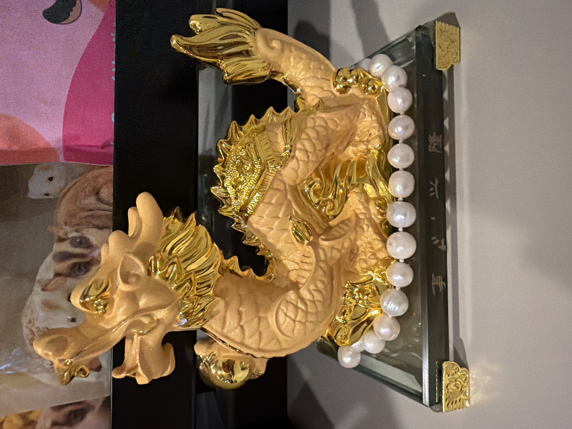 The image displays a golden dragon sculpture, oriented vertically, mounted on a clear glass or acrylic plaque. The dragon features a detailed design with a matte gold body, enhanced by glossy, reflective gold accents on its scales, mane, claws, and facial features. A strand of lustrous, irregularly shaped white pearls is draped across the dragon's midsection.
The clear plaque has decorative gold-colored corner pieces at its top and bottom. Vertical script, likely Chinese characters, is inscribed on the plaque behind the pearls. The uppermost visible character is clearly "龍" (lóng), meaning dragon, followed by other less distinct characters, possibly forming a phrase such as "龍飛...年" (Dragon flying... year).
The background on the left side of the image shows a vibrant, patterned surface, primarily pink and red, with embedded images of several cats or kittens. This is adjacent to a solid black surface, which runs along the left edge of the dragon display. The setting appears to be indoors, possibly on a shelf or against a wall. The entire image is rotated 90 degrees counter-clockwise from what might be its natural display orientation. There are no people visible, and no activity is taking place; it is a static display of an ornate cultural or decorative item.