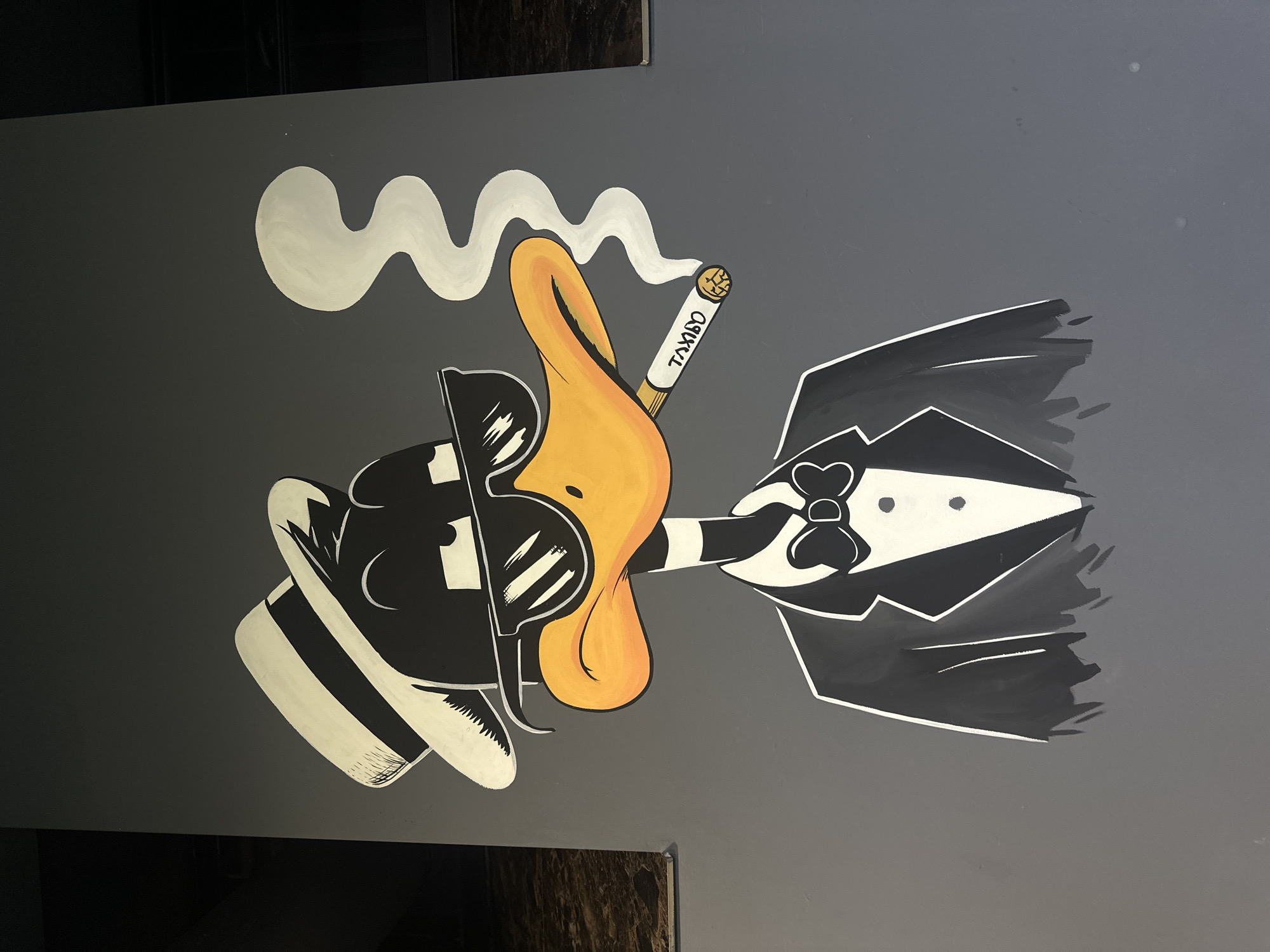 The image depicts a cartoon mural of Daffy Duck in Yerevan, Armenia. Daffy is portrayed in a sophisticated manner, wearing a black suit with a white bowtie, a white-striped black fedora, and sunglasses. He is holding a cigarette in his beak that reads "TAXBO." A trail of white smoke emanates from the cigarette. The mural is painted on a grey wall.