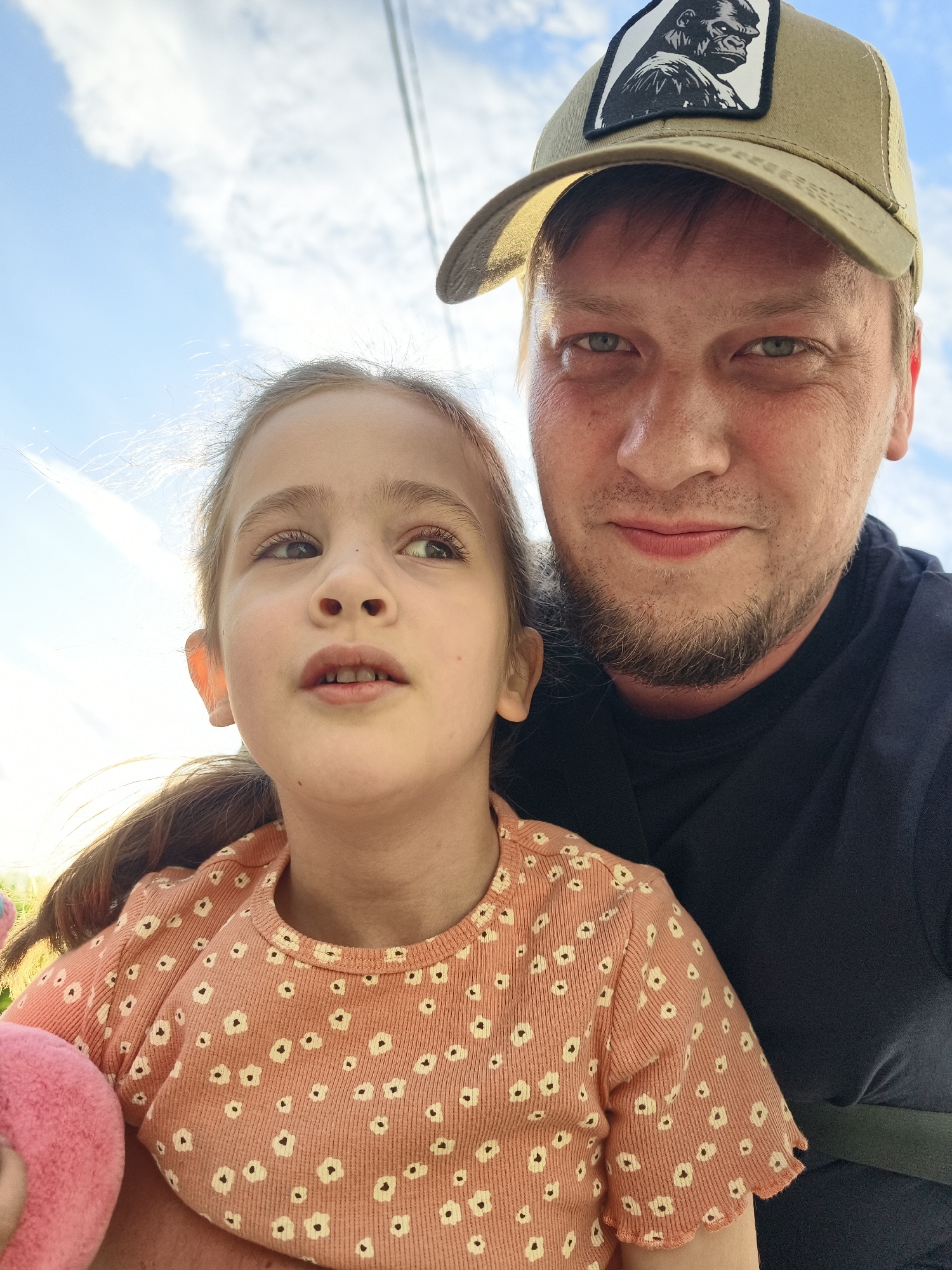 A selfie shows a man and a girl outdoors. The man wears a tan baseball cap with a gorilla patch, and a dark-colored t-shirt. He has fair skin, light eyes, and a beard. The girl has fair skin, brown hair in a ponytail, and an orange shirt with white floral pattern. Both are looking slightly upward. The background shows a cloudy sky and electrical wires. It is possible that the image was taken in Chaikovsky, Russia, although no specific indicators from the photo confirm this.