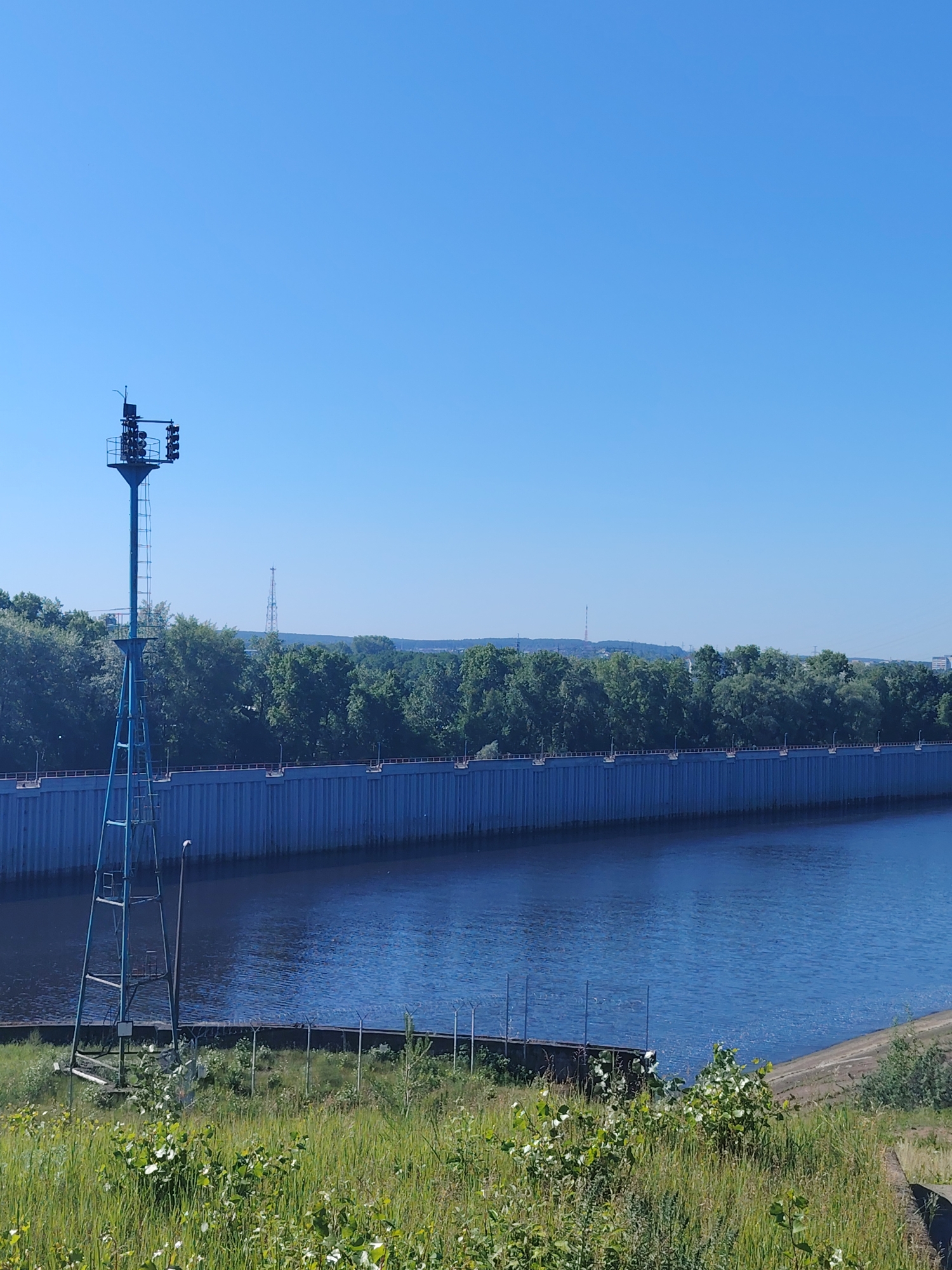 The image depicts a wide blue river alongside a tall blue metal wall, likely part of a dam or flood control system. On the left, a blue metal tower with multiple signal lights stands prominently. Behind the wall, a dense line of green trees stretches across the landscape, with a few distant cell towers visible on the horizon under a clear blue sky. The foreground is covered in lush green grass and various low-lying plants. This scene is located in Chaikovsky, Russia.