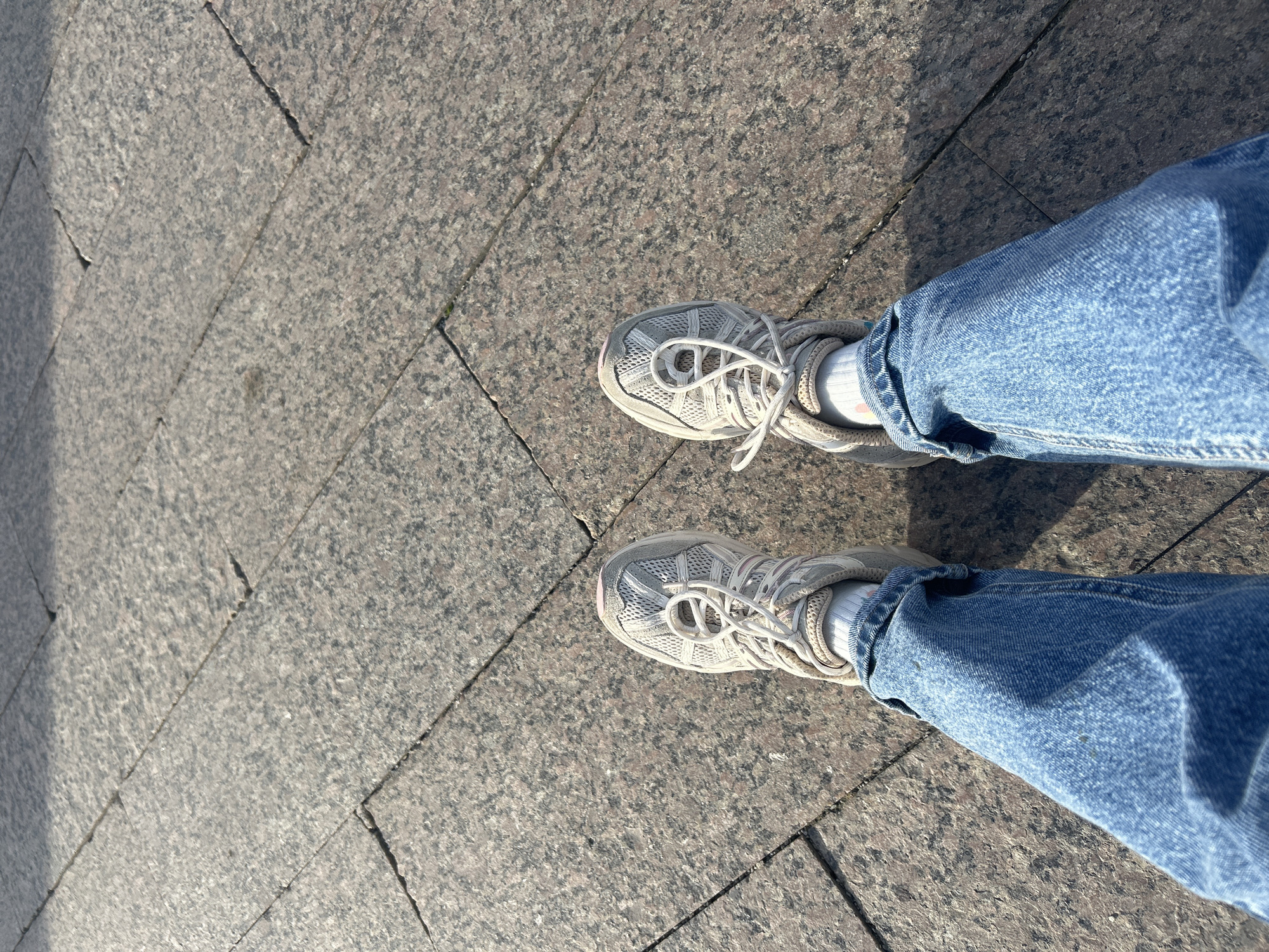 An overhead shot captures a person's feet standing on a sunlit paved surface, typical of an urban environment in Moscow, Russia. The ground is composed of large, rectangular stone tiles, likely granite, exhibiting a speckled pattern of various shades of grey and reddish-brown, with visible grout lines between them.
The subject is wearing light-wash denim jeans, which are cuffed or rolled up just above the ankles. Beneath the jeans, white socks are visible, with a subtle pink detail or mark on one of them. The person is wearing light grey or white athletic shoes, commonly known as sneakers, with prominent white laces tied on top. The shoes feature a mesh-like upper material and a somewhat thick sole, with a slightly darker, possibly pale pink or beige, accent near the toe cap.
Strong natural sunlight illuminates the scene from the right, casting distinct, sharp shadows across the left side of the ground and partially obscuring the left leg and shoe. This suggests it is a bright, clear day. The activity depicted is simply standing or walking, captured from a first-person or top-down perspective focusing on the footwear and ground. No other subjects or text are visible in the image.
