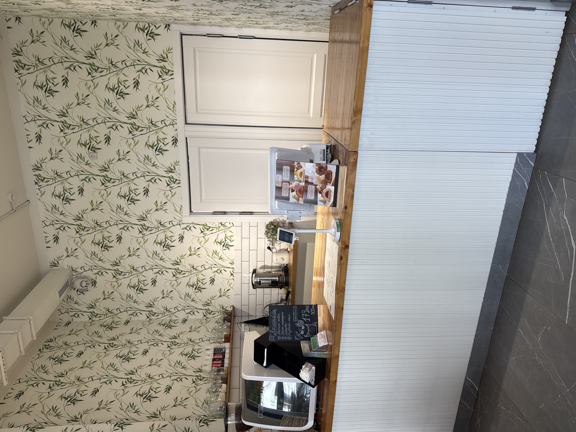 A high-angle, slightly tilted shot shows the interior of a cafe or shop with a modern and nature-inspired design. The walls and ceiling are covered in a white wallpaper with a repeating pattern of green bamboo stalks and leaves. A wooden counter runs across the foreground, with a white, ribbed wall paneling behind it. On the counter, a digital menu display shows images of pastries. To the left of the menu, a small chalkboard sign is visible with handwritten text in Russian. Among the visible text are the words "Лимонады" (Lemonades) and "Моти" (Mochi), along with prices like "179 руб" (179 rubles). Behind the counter, there are shelves with glass jars containing various goods, possibly coffee beans or spices. A coffee machine and other kitchen appliances are also visible. The floor in the lower right corner of the image is made of dark gray tiles with white veining. The scene appears to be indoors, with artificial lighting. No people are visible in the frame. The overall impression is of a clean, stylish, and possibly upscale establishment.