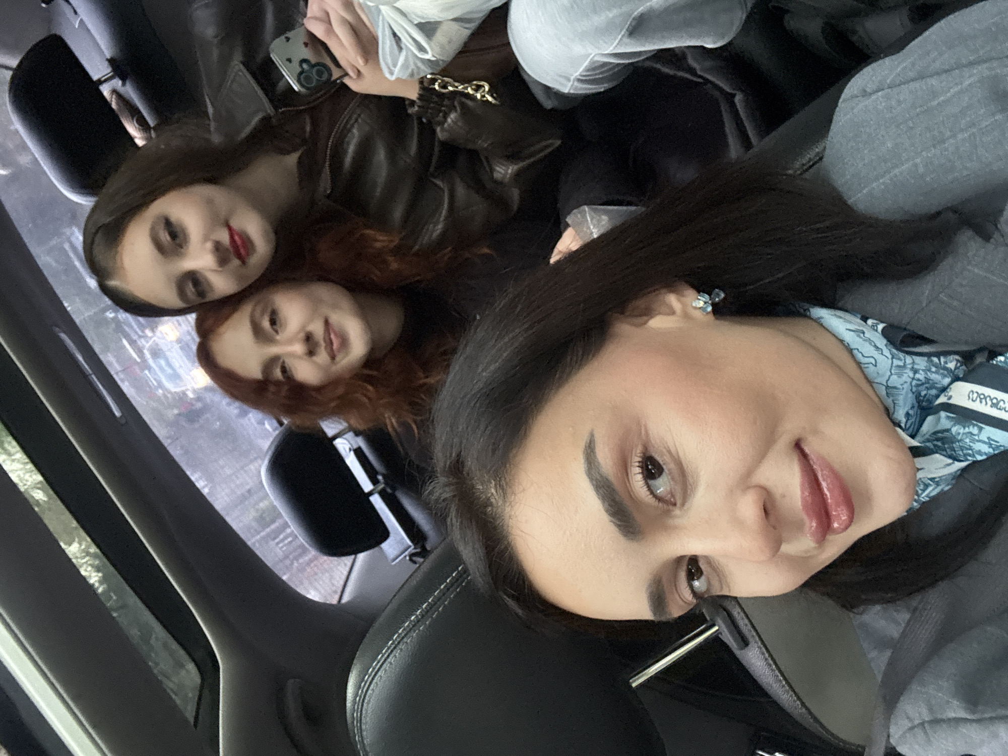Three women are in the back of a car. One has long dark hair and is wearing a grey jacket and blue patterned scarf. Another woman has red hair. The third woman has dark hair and is wearing a brown leather jacket. One is holding a phone in a case with a heart design. The background through the car window shows a city street, possibly in Tbilisi, Georgia.
