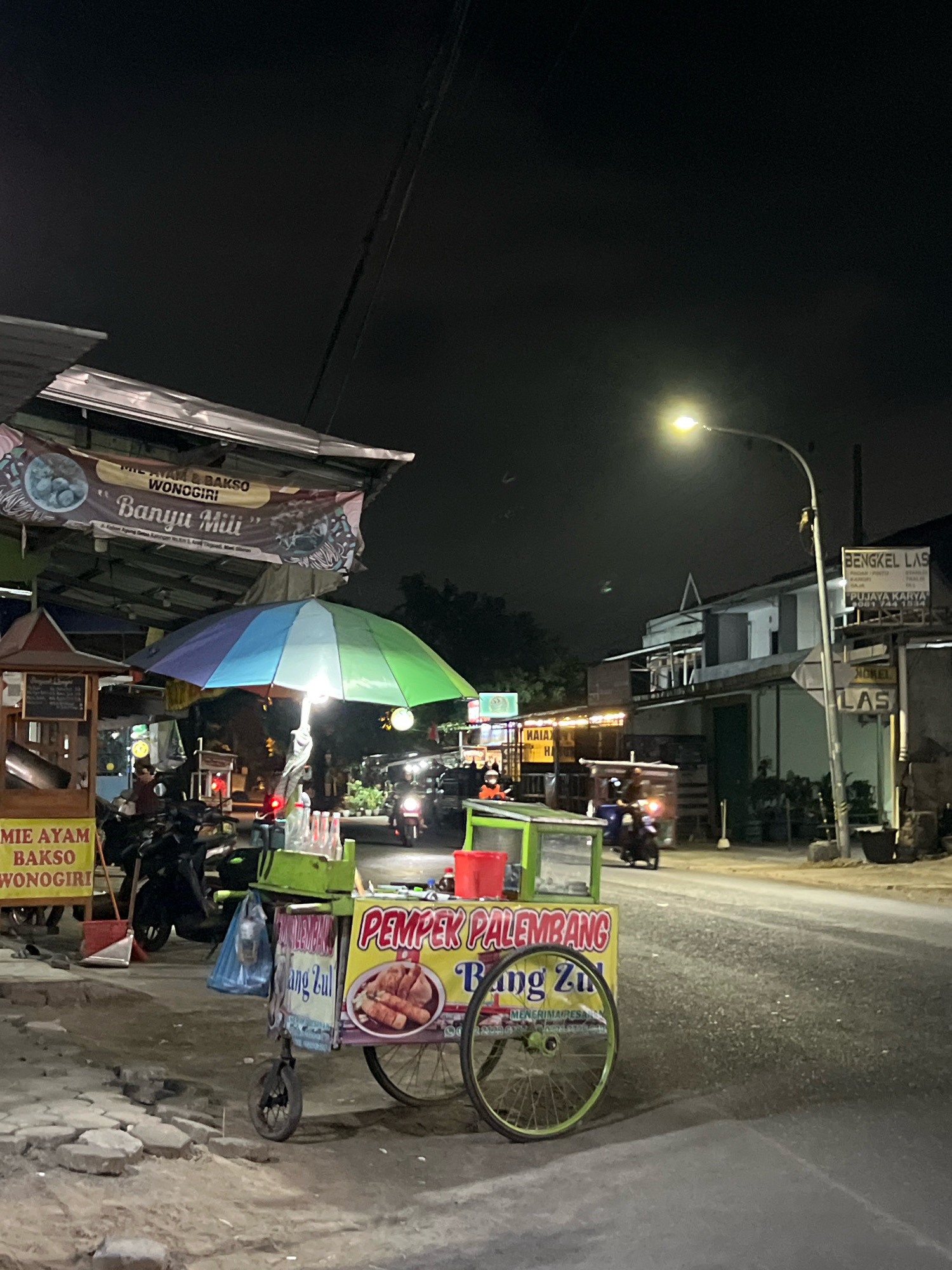 The image depicts a nighttime street scene in Tlogoadi, Indonesia, likely a local market or food stall area. In the foreground, a mobile food cart selling "Pempek Palembang Bang Zul" is prominently displayed. The cart has a yellow body with red lettering and features a glass display case. Next to it, another cart sells "Mie Ayam & Bakso Wonogiri" under a banner with the name "Banyu Mili". A colorful, multi-panel umbrella provides shade over the food cart area. Several motorcycles are parked and visible in the background, with some people present on them. The street is paved and appears wet in places, reflecting the light from a streetlamp. Buildings line the street, one of which has a sign for "BENGKEL LAS" (Welding Shop) and "HOTEL LAS". The overall atmosphere is that of a bustling, casual nighttime eatery.