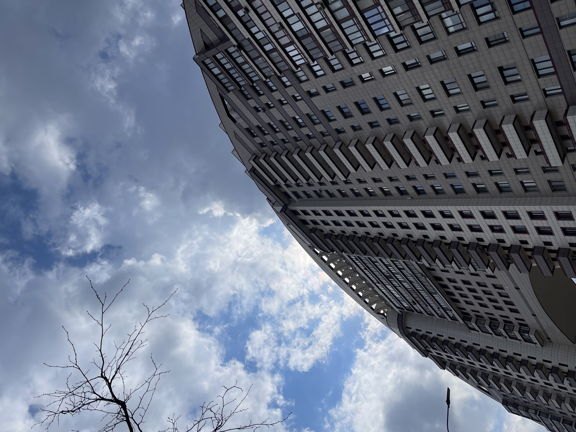 An upward angle captures the building "House on Mosfilmovskaya Street" in Moscow, Russia. A portion of the building takes up the upper right, its gray facade dotted with evenly spaced windows and projecting rectangular balconies. To the left and in the background, a sky of interspersed blue and white clouds is visible. The bare branches of a tree are noticeable in the lower left corner.
