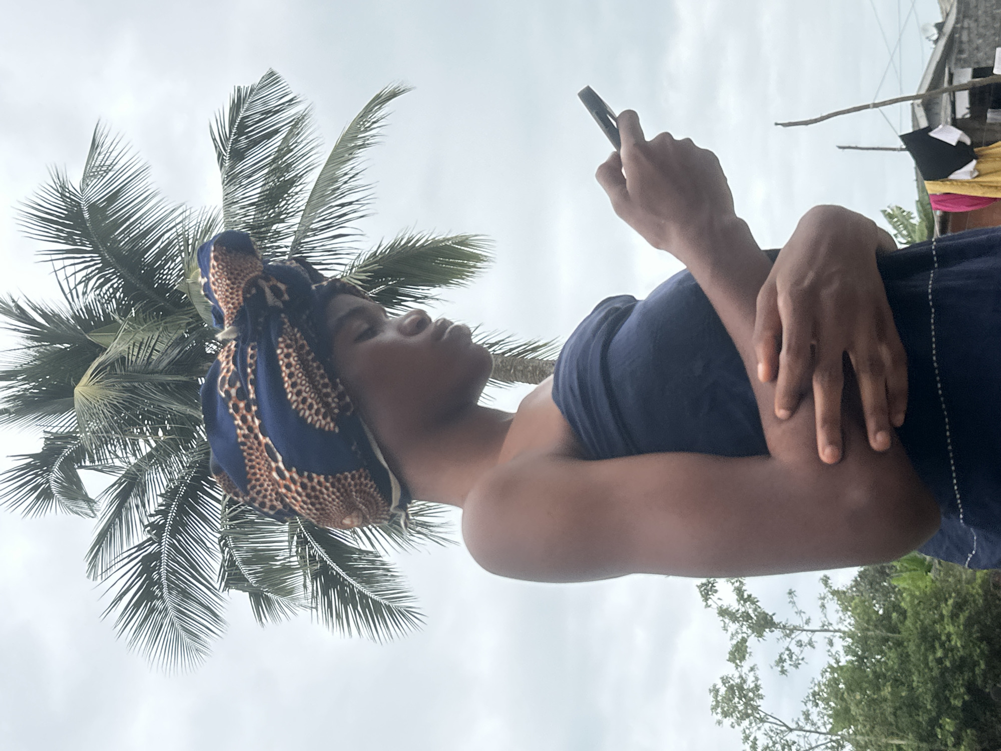 A young woman with dark skin, wearing a dark blue strapless top and a patterned headwrap, stands outdoors. She is looking down at a black mobile phone she holds in her right hand, with her left arm resting across her midriff. Behind her, a large palm tree with green fronds extends across the upper portion of the frame against a cloudy sky. In the background to the left, lush green foliage is visible, and to the right, a clothesline with several garments hangs between two wooden poles in front of a modest structure. This scene is set in the city of Badagry, Nigeria.