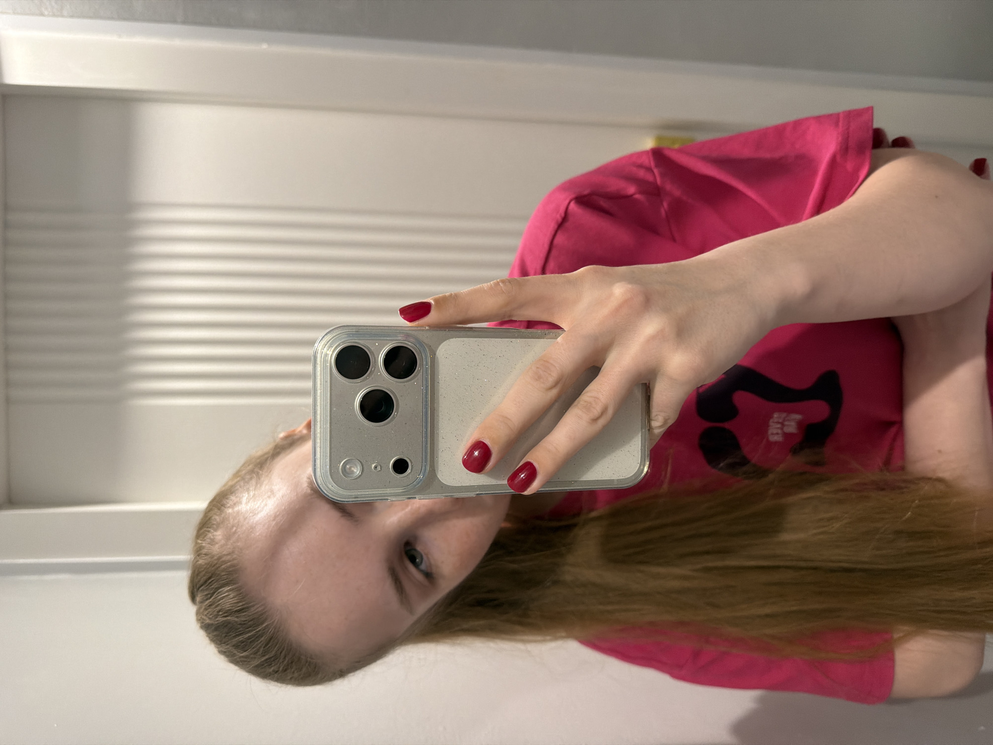A person is holding a mobile phone in their right hand, taking a selfie. The person has blonde hair and is wearing a pink shirt. The background shows a white door with horizontal grooves. The location appears to be indoors.