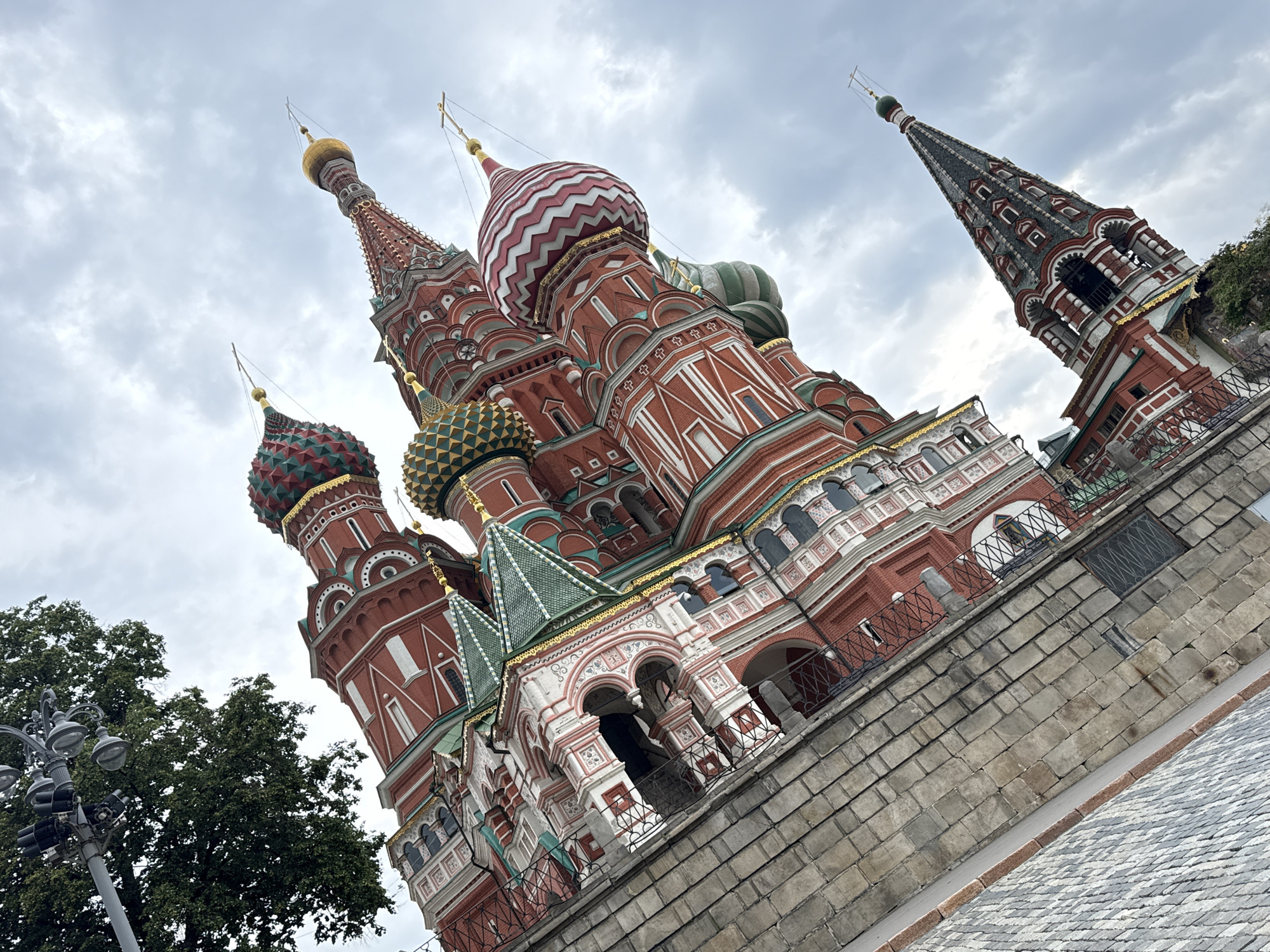 The image shows a low-angle, tilted view of St. Basil's Cathedral in Moscow, Russia. The iconic onion domes, red brick walls with white detailing, and green tiled roofs are prominent. The sky is overcast with clouds. A portion of a cobblestone ground is visible in the foreground, along with some greenery and a lamppost in the upper left corner. The cathedral is a historic landmark located on Red Square in Moscow.