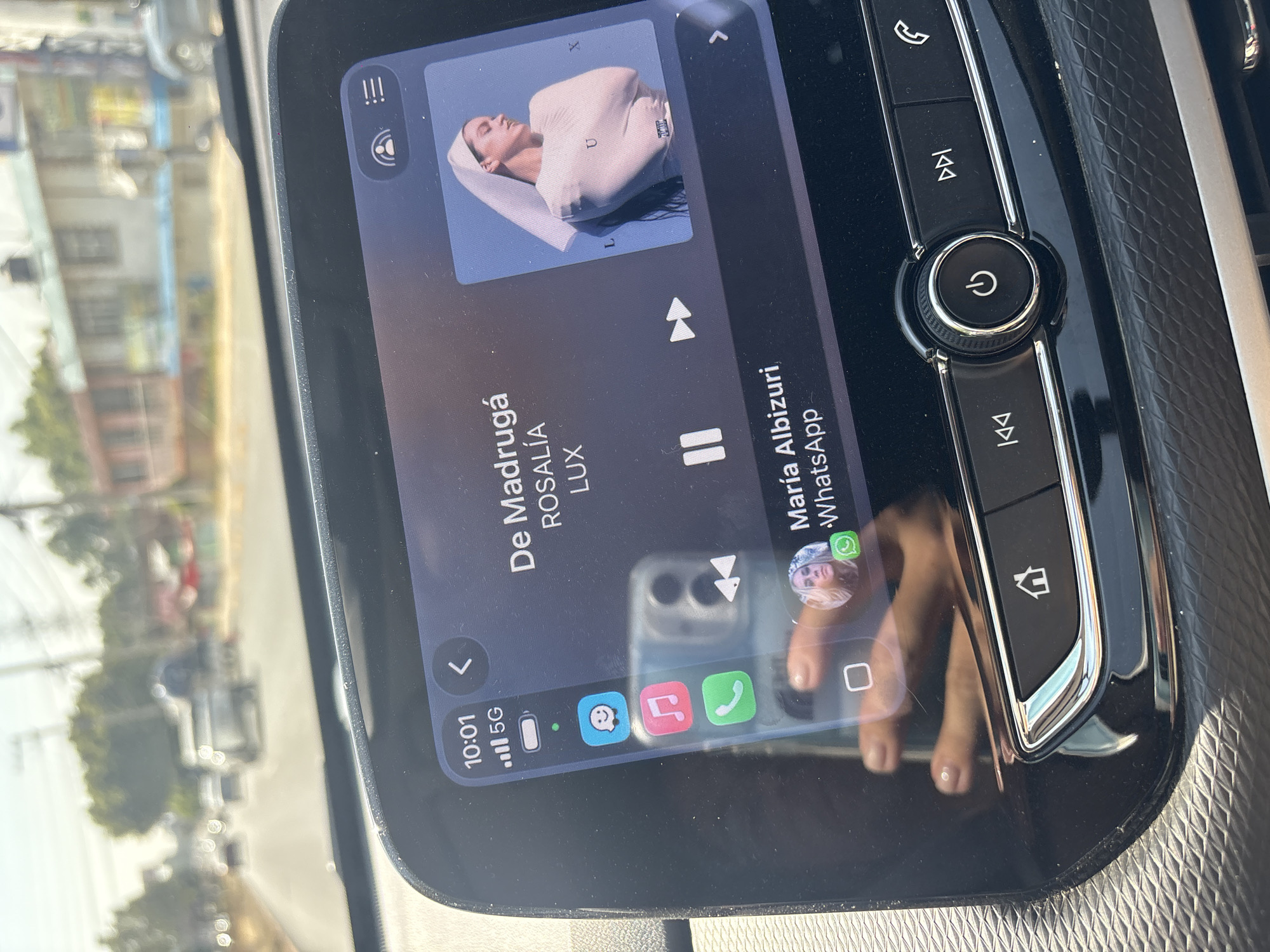The image depicts the interior of a vehicle, specifically focusing on its infotainment screen and the external environment through the windshield.
The infotainment screen displays an active music player interface at 10:01 AM, showing a strong 5G cellular signal and a partially charged battery. The song "De Madrugá" by Rosalía, from the album "LUX," is featured, with album art portraying a woman draped in light-colored fabric. Application icons for Snapchat, Music, and Phone are visible on the left side of the screen. A WhatsApp notification for "María Albizuri," including a profile picture, appears below the music track information. A clear reflection on the screen shows a hand holding a light-colored smartphone, likely used to capture the image, with visible fingers exhibiting manicured nails. Below the screen, physical vehicle controls are present, including a chrome-trimmed "home" button, track skip buttons, and a central rotary knob marked with a power/mute symbol. The dashboard material around these controls features a textured, diamond-patterned finish.
Through the windshield, a multi-lane asphalt road is visible under bright daylight conditions. Several vehicles, including cars and a truck, are present in the distance, indicating traffic flow. On the right side of the road, various buildings of different architectural styles, some with balconies and painted in light colors, are interspersed with trees and utility poles.
No specific landmarks, unique architectural features, or discernible street signage within the image allow for precise geographical identification within Heroica Puebla de Zaragoza, Mexico. The scene represents a typical urban or suburban road environment.