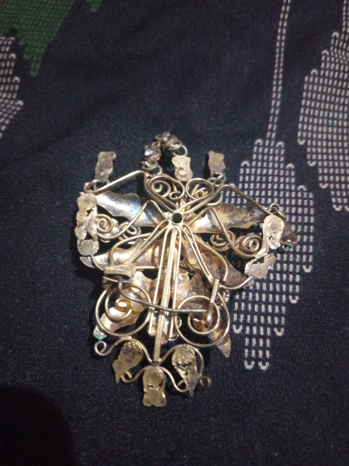 The image, captured in Hasanpur, India, displays an overhead close-up view of the reverse side of an ornate metallic object, consistent with aged jewelry or an ornament. The object features intricate filigree wirework, including curved scrolls, stylized leaf-like shapes, and a central structural element. Multiple small loops and prongs are visible along the perimeter and on the upper edge, indicating attachment points. A pin-back clasp mechanism is partially visible on the reverse. The metallic surface exhibits irregular dark tarnishing and localized greenish oxidation. The object rests on a dark fabric surface patterned with repeating light-colored geometric shapes, primarily small, interconnected rectangular or square grids. No individuals or dynamic actions are depicted.