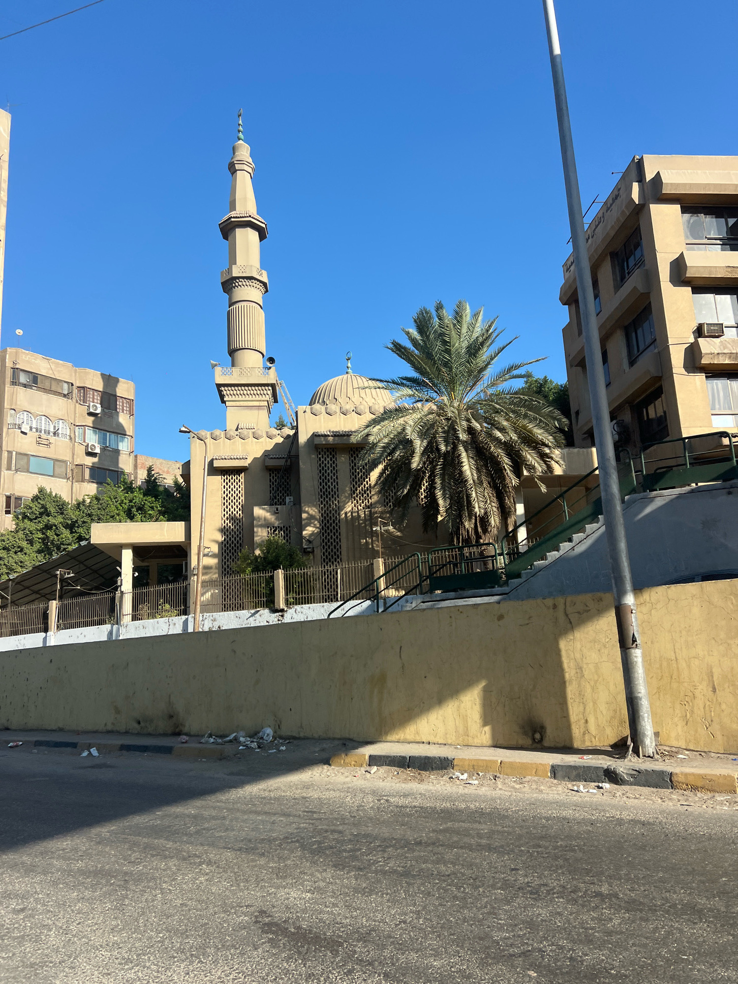 A mosque with a tall minaret is visible in Giza, Egypt, under a clear blue sky. The mosque is made of tan-colored stone and features a dome and arched windows. A large palm tree stands in front of the mosque. Adjacent to the mosque are several multi-story buildings, also constructed with tan-colored materials. A street runs in the foreground, with some litter scattered along the curb. A tall, thin metal pole, possibly a lamppost or utility pole, is situated in the middle ground, casting a shadow on the street and wall. The overall scene depicts an urban environment with religious and residential structures.