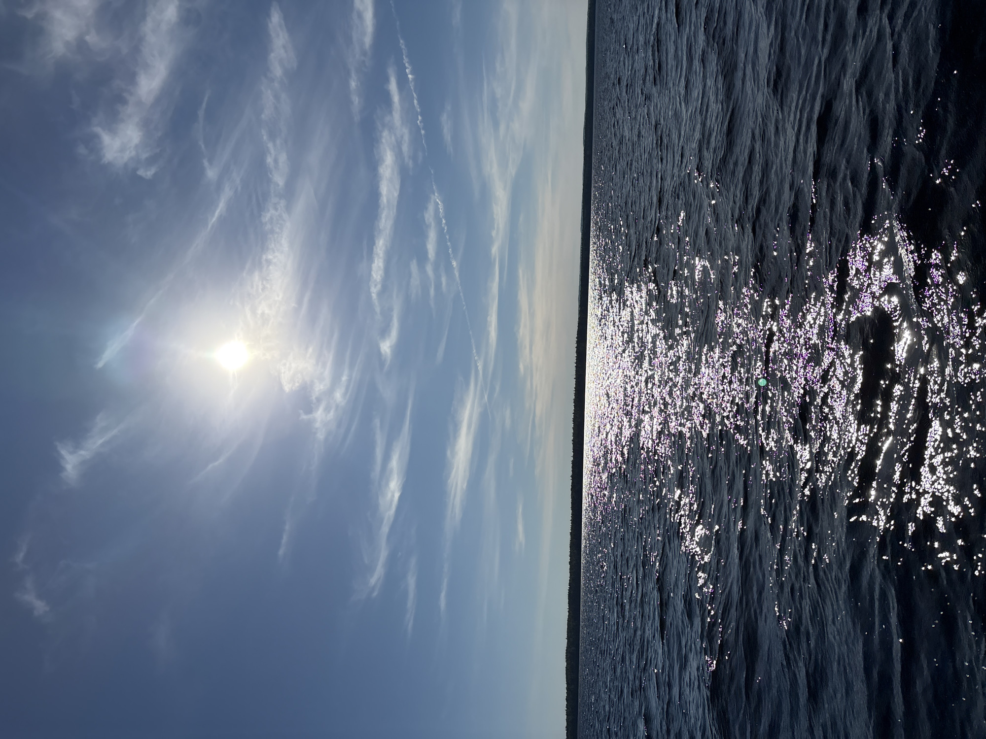 A wide-angle, landscape view captures a sunlit expanse of dark blue water. The sun, positioned in the upper center of the frame, is bright and slightly obscured by wispy white clouds that stretch across the clear blue sky. A band of dark green land, densely forested, forms a horizon line across the middle of the image. The water's surface is disturbed by small waves, and the sun's reflection creates a shimmering, sparkling path of light extending from the lower center towards the horizon. The scene is likely a natural outdoor setting, potentially a lake or large body of water, given the presence of the forest and the vastness of the water. There are no people or man-made structures clearly visible in the image.